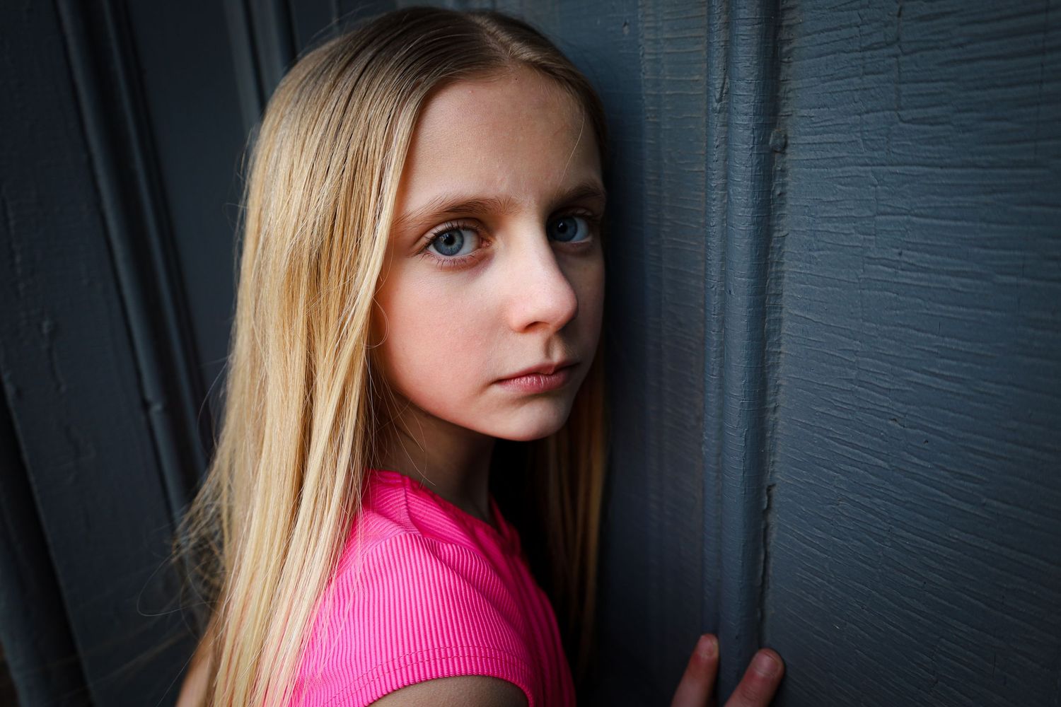 Close-up portrait of a blonde girl with blue eyes leaning against a dark wooden wall.