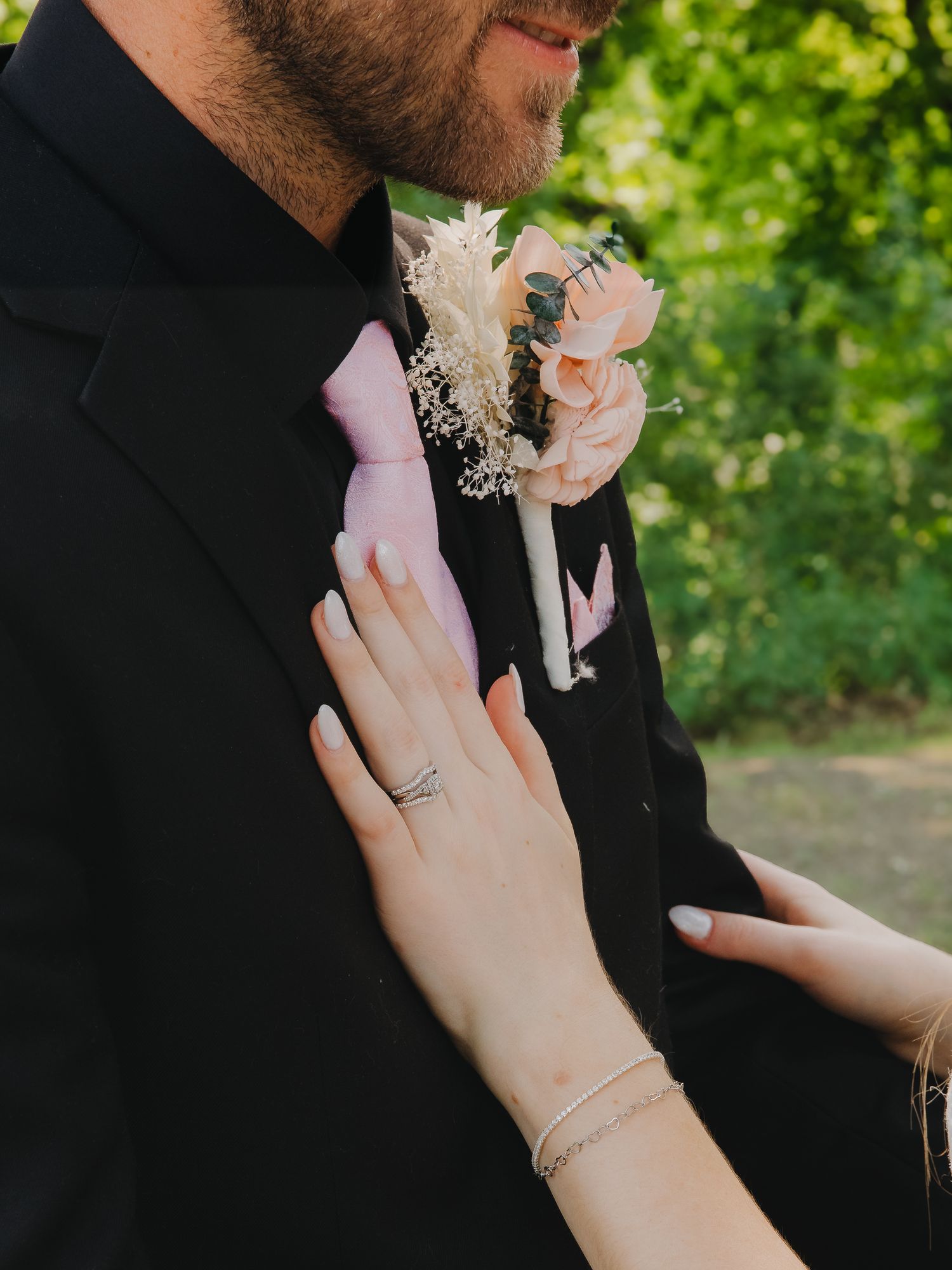 Bride adjusts grooms pink rose boutonniere on black suit jacket outdoors.