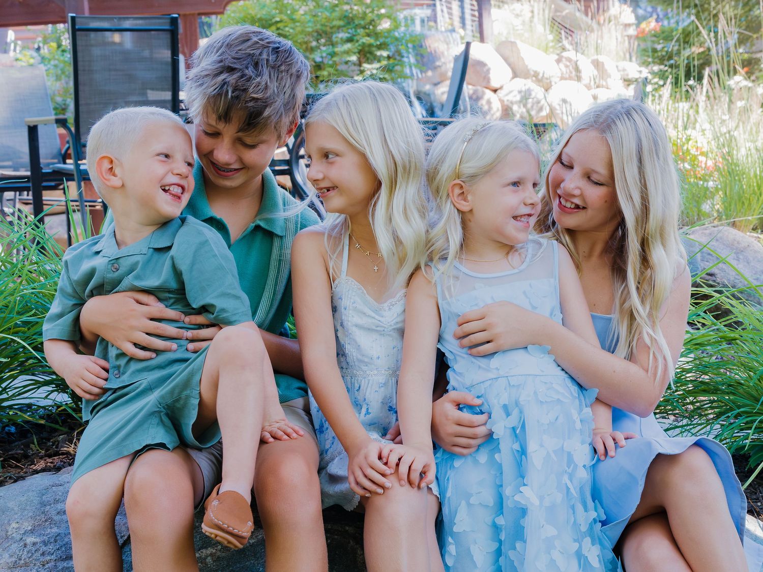Five siblings laugh and embrace while seated together in a lush outdoor garden setting.