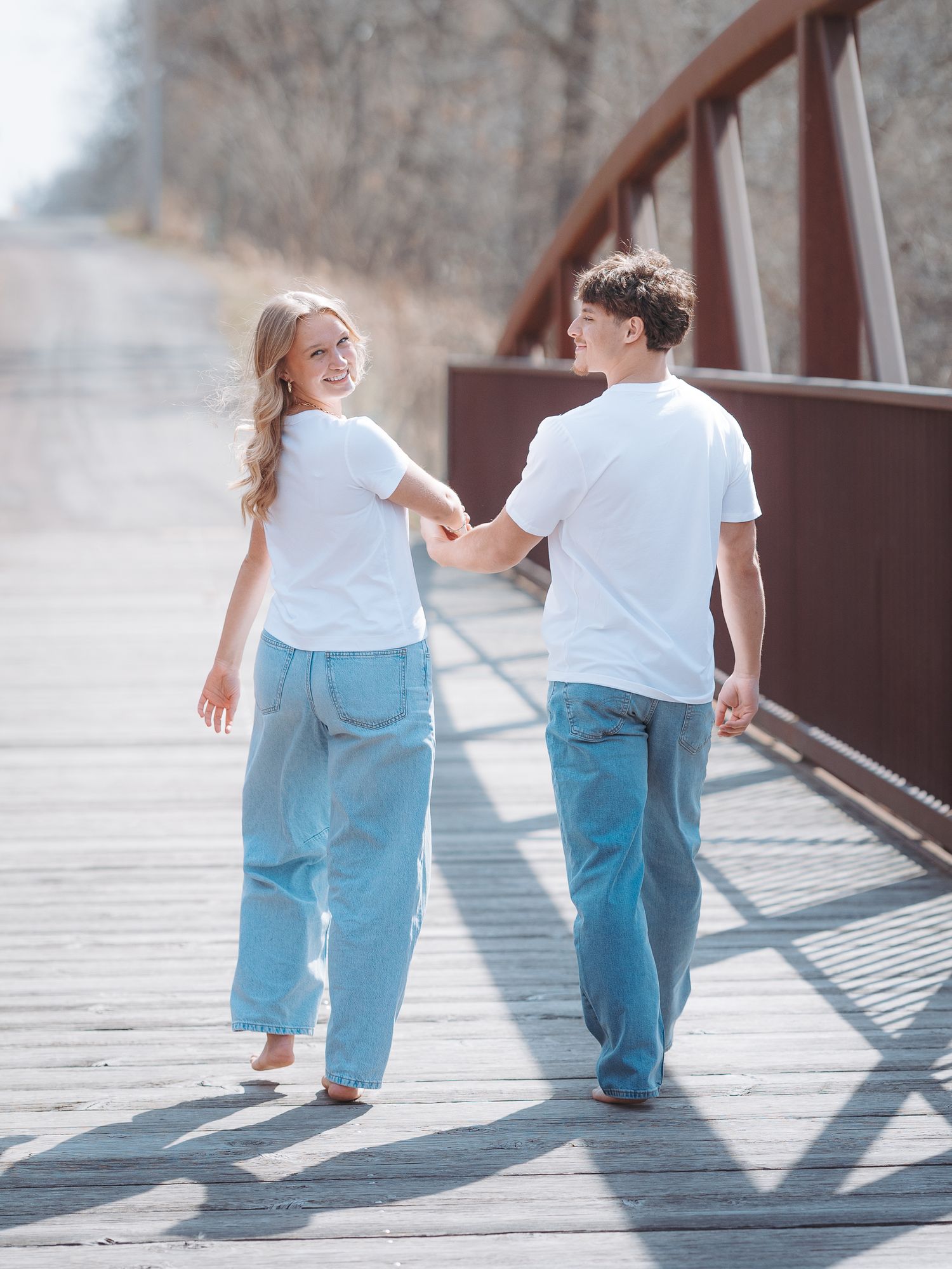 Young couple holding hands walking on a wooden bridge, wearing matching white tees and light blue jeans.