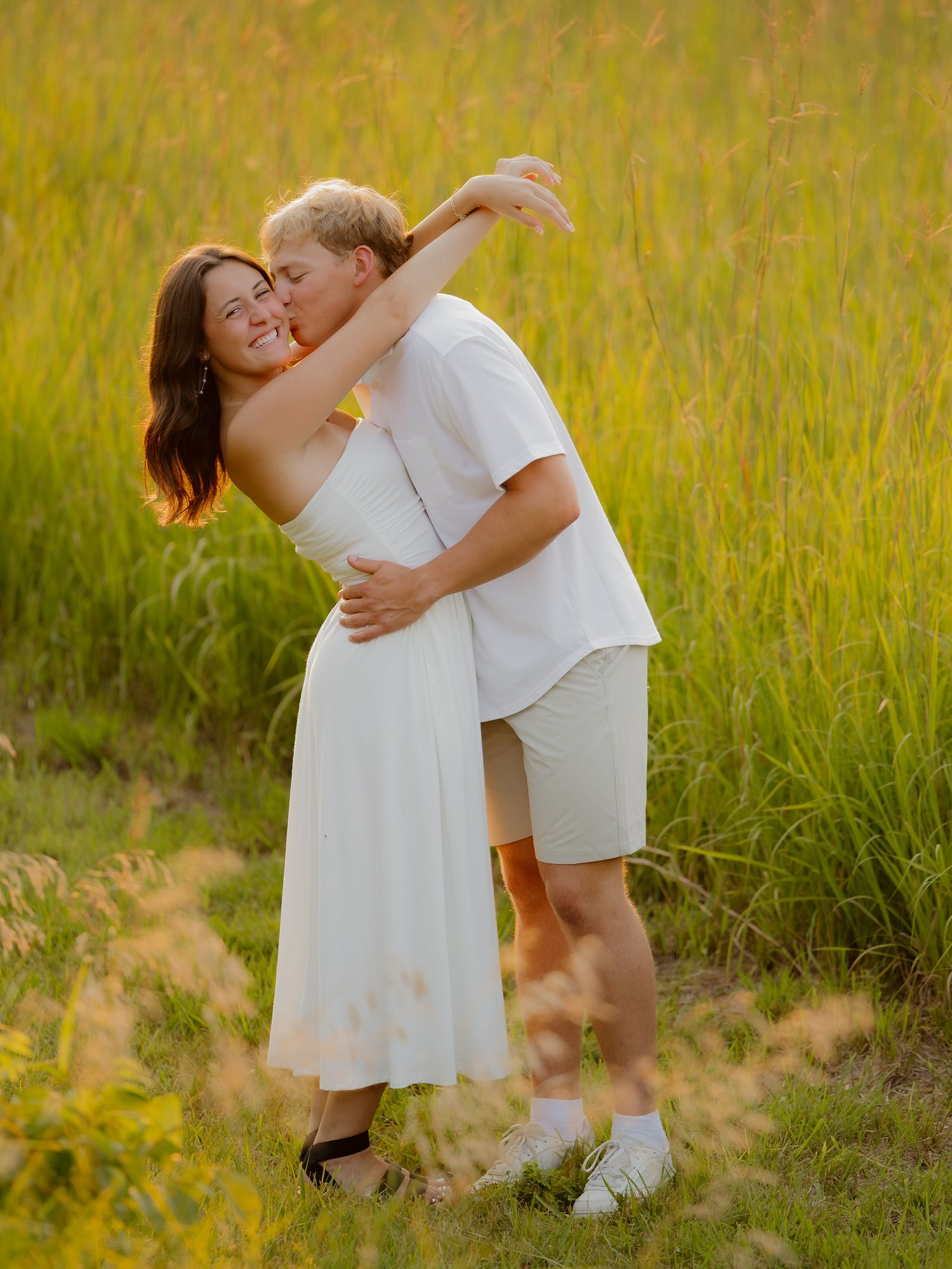 Couple embracing in golden sunlit field, woman in white dress smiling as man kisses her cheek.