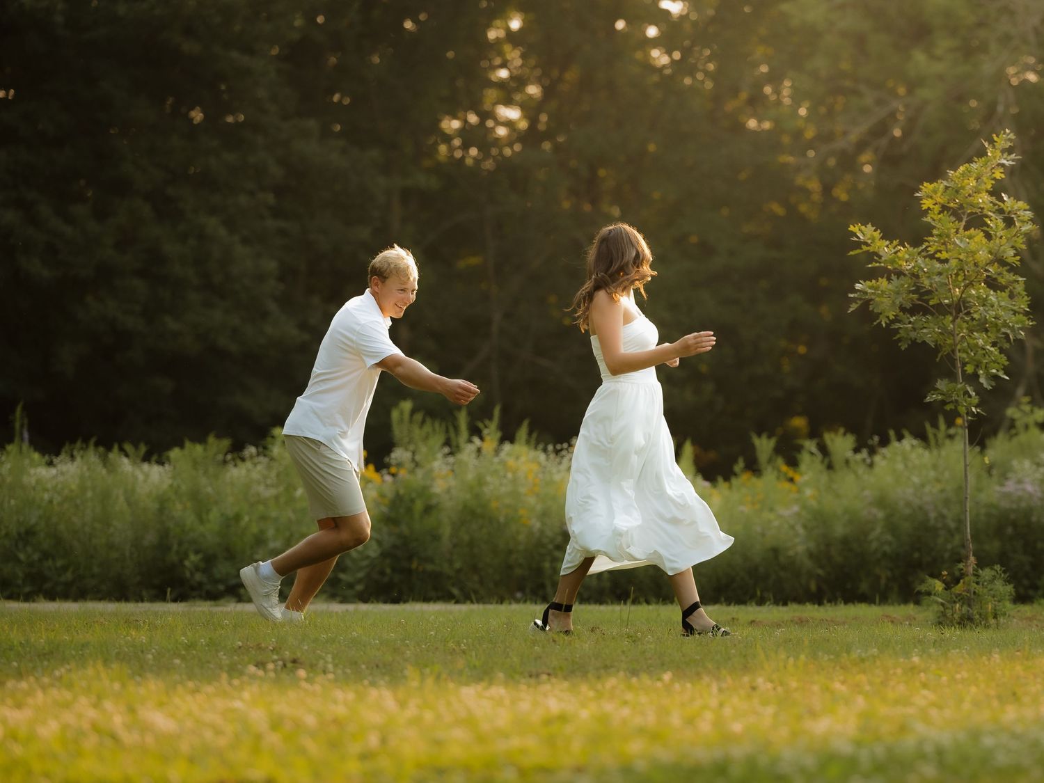 Couple in white outfits running playfully through a sunlit green park surrounded by lush trees.
