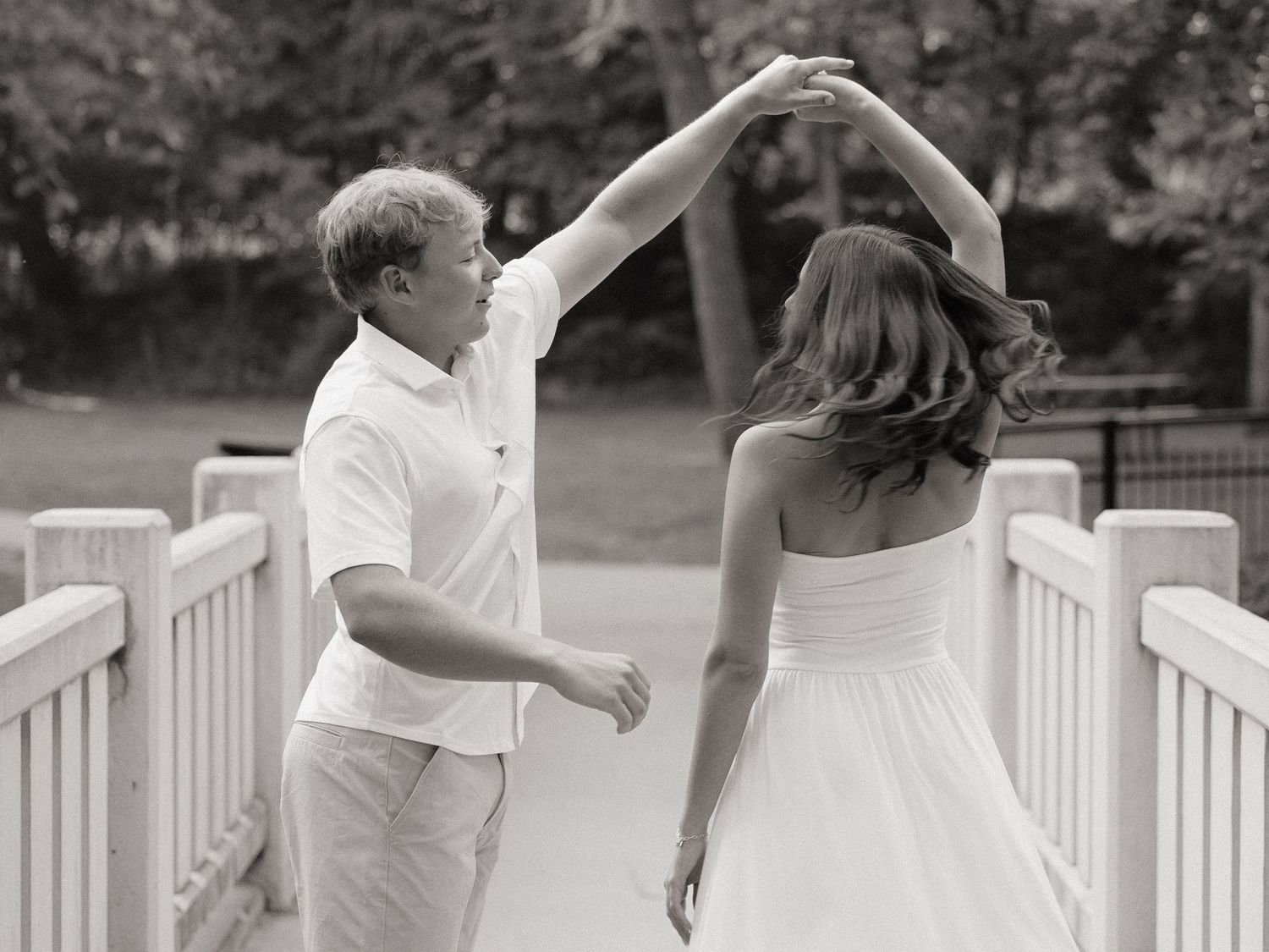 Black and white photo of a couple dancing on a bridge, man spinning woman in a white dress outdoors.