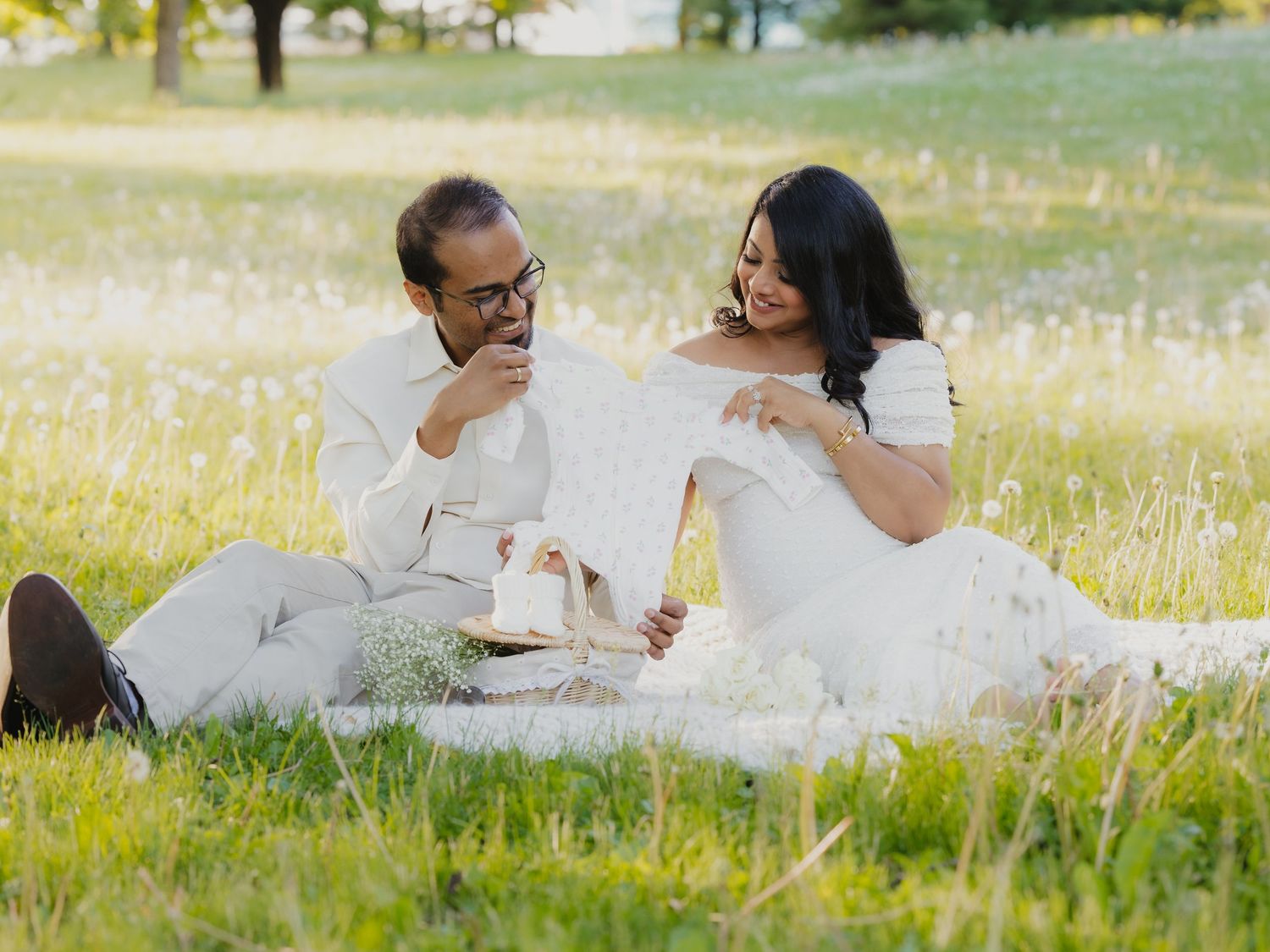 Couple in white sitting in sunlit meadow laughing together over a picnic basket.