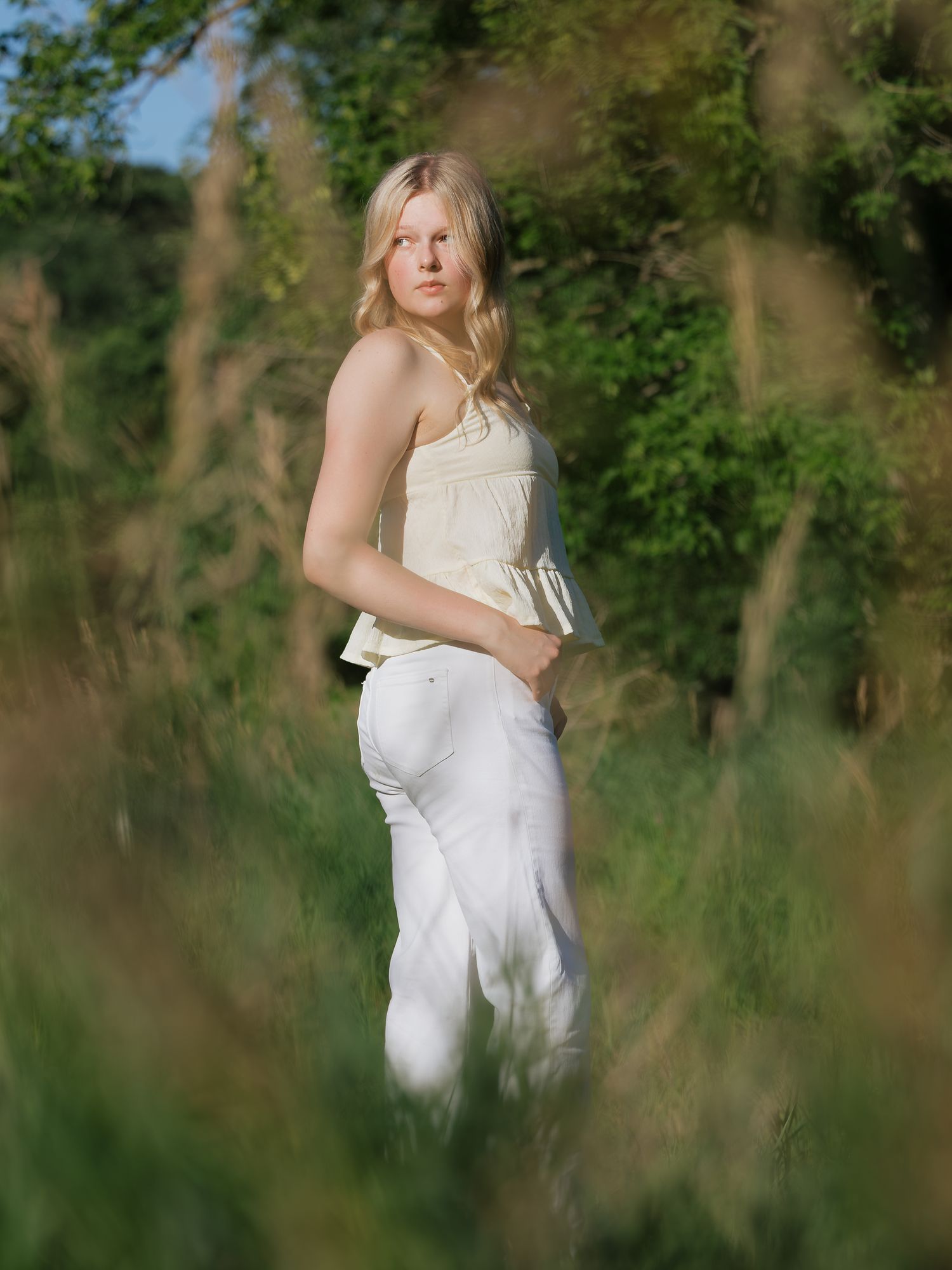 Blonde woman in white jeans and yellow camisole standing in a lush green meadow.
