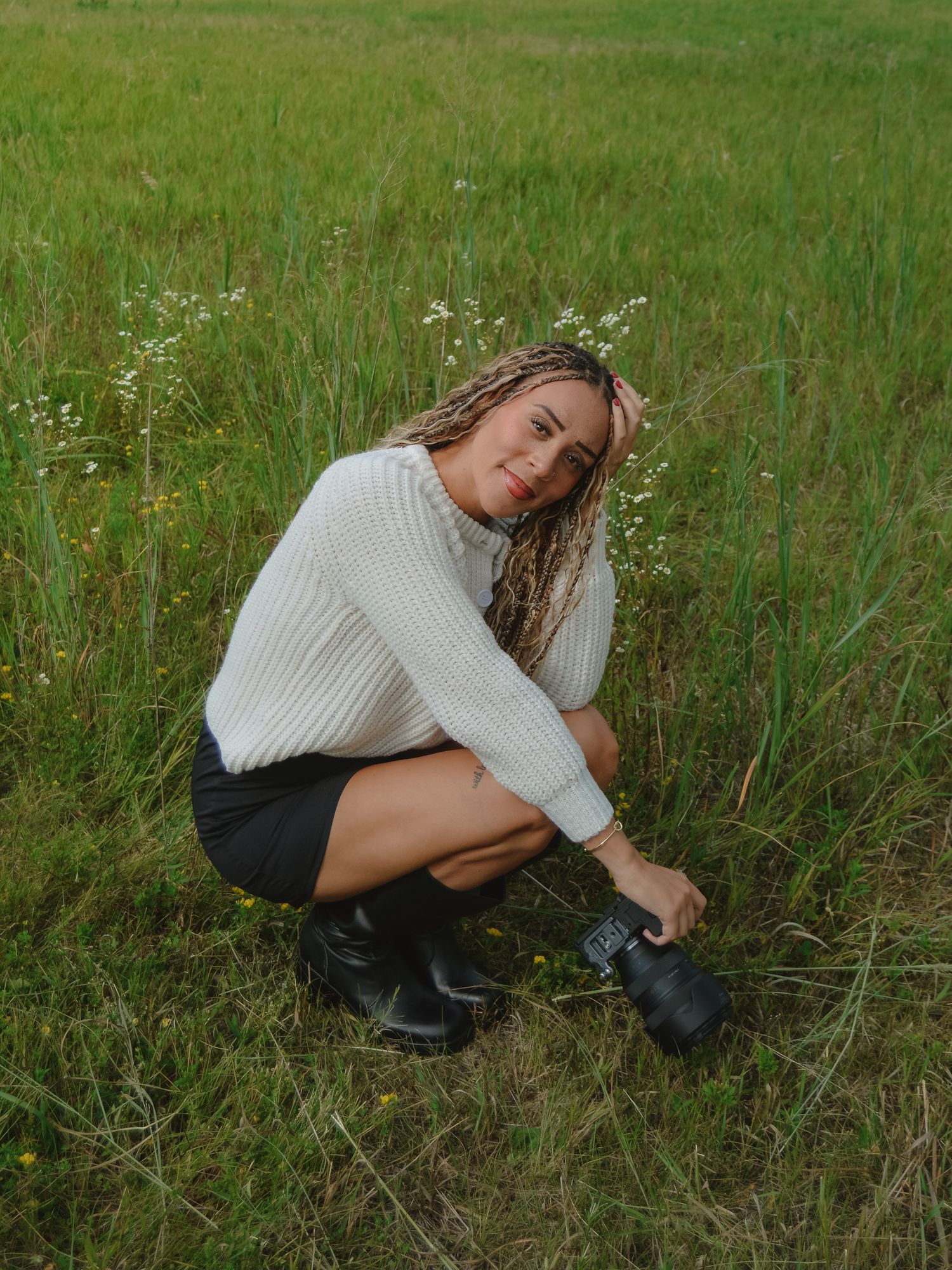 Young woman crouching in a green field, wearing a cream sweater and black skirt, holding a camera.