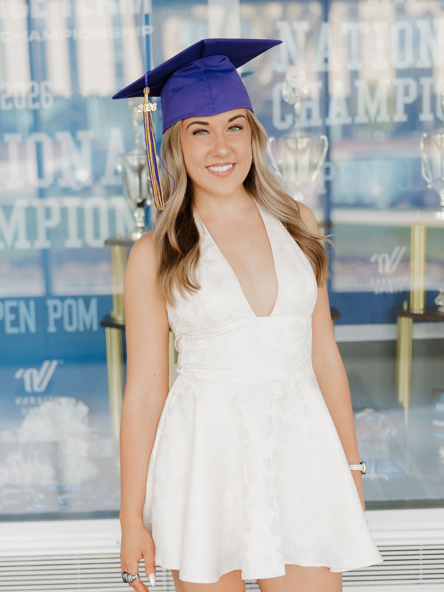 Smiling graduate in white dress and blue cap poses in front of National Champions display.