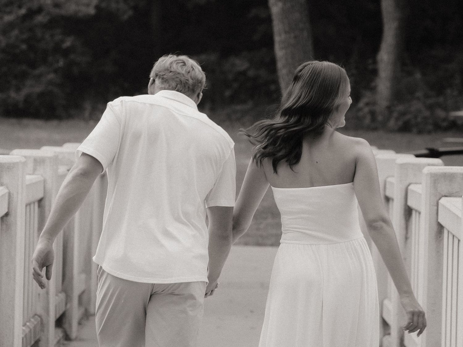 Black and white photo of a couple holding hands walking on a bridge, woman in white dress, man in white shirt.