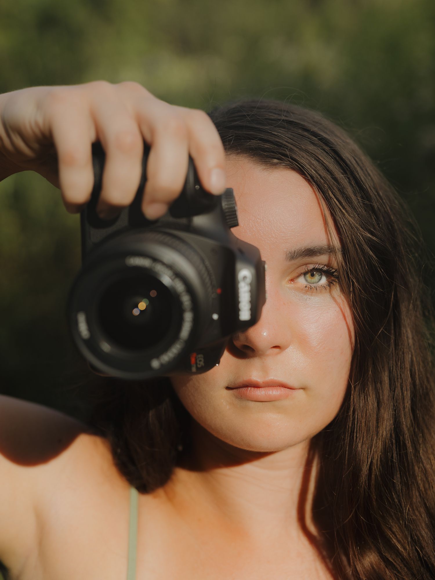 Young woman holding a Canon DSLR camera up to her eye outdoors in natural light.