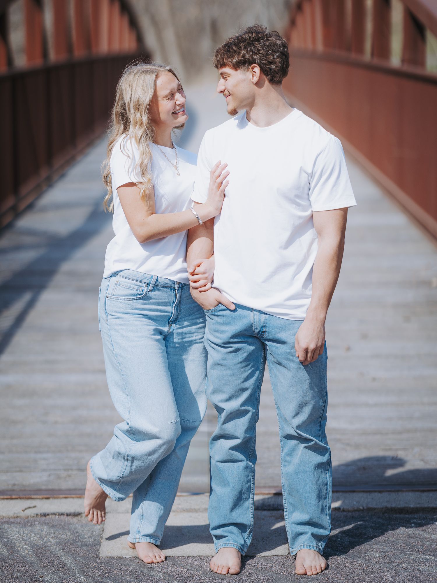 Couple in matching white tees and light jeans smiling at each other on a rustic red bridge.