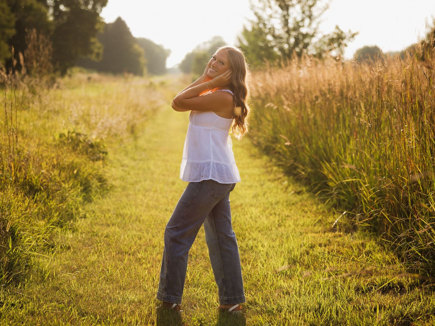 Young woman in white top and jeans standing on a sunlit meadow path, embracing nature in golden hour light.