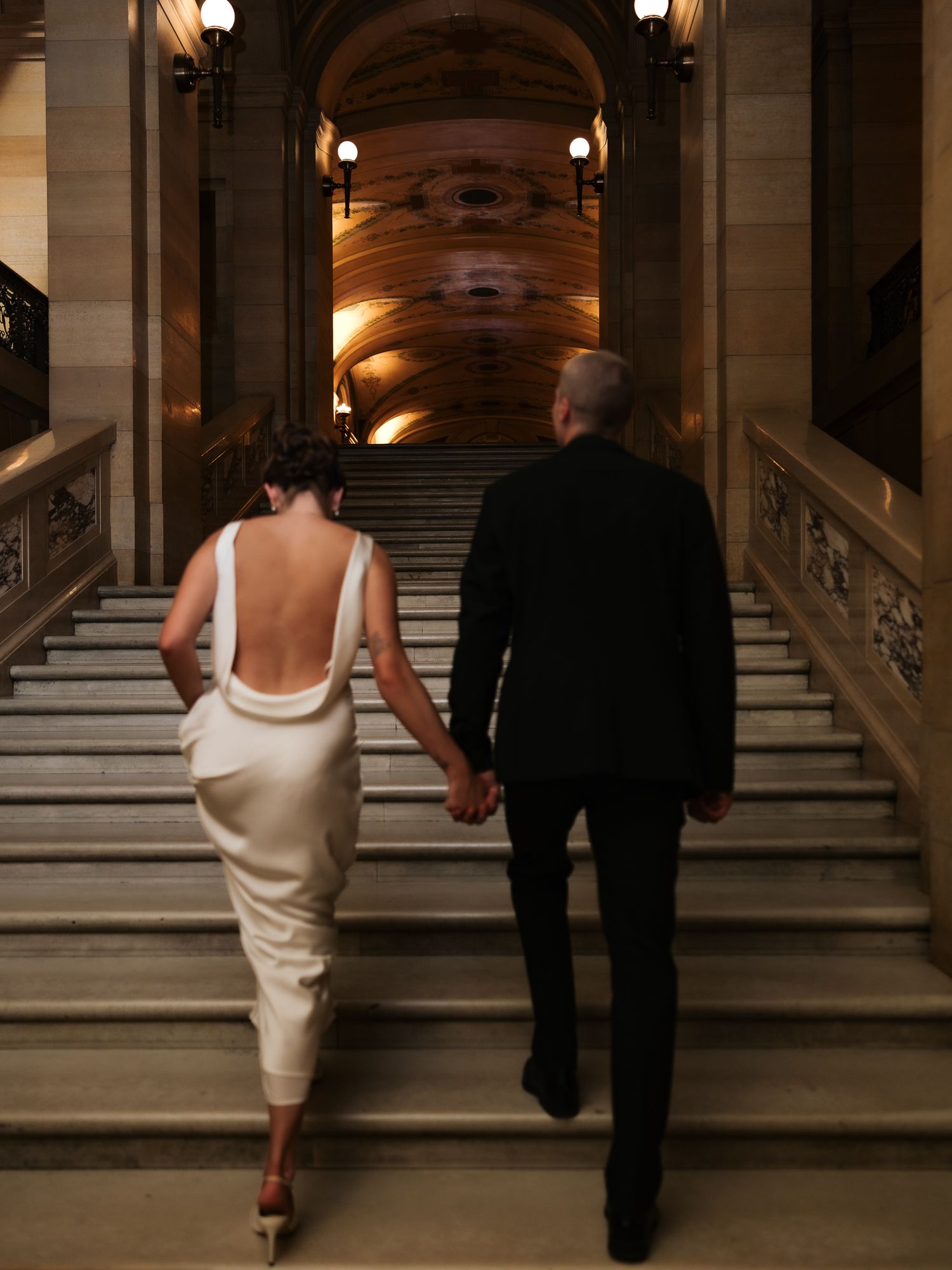 Bride and groom holding hands walking up grand staircase in elegant historic building.