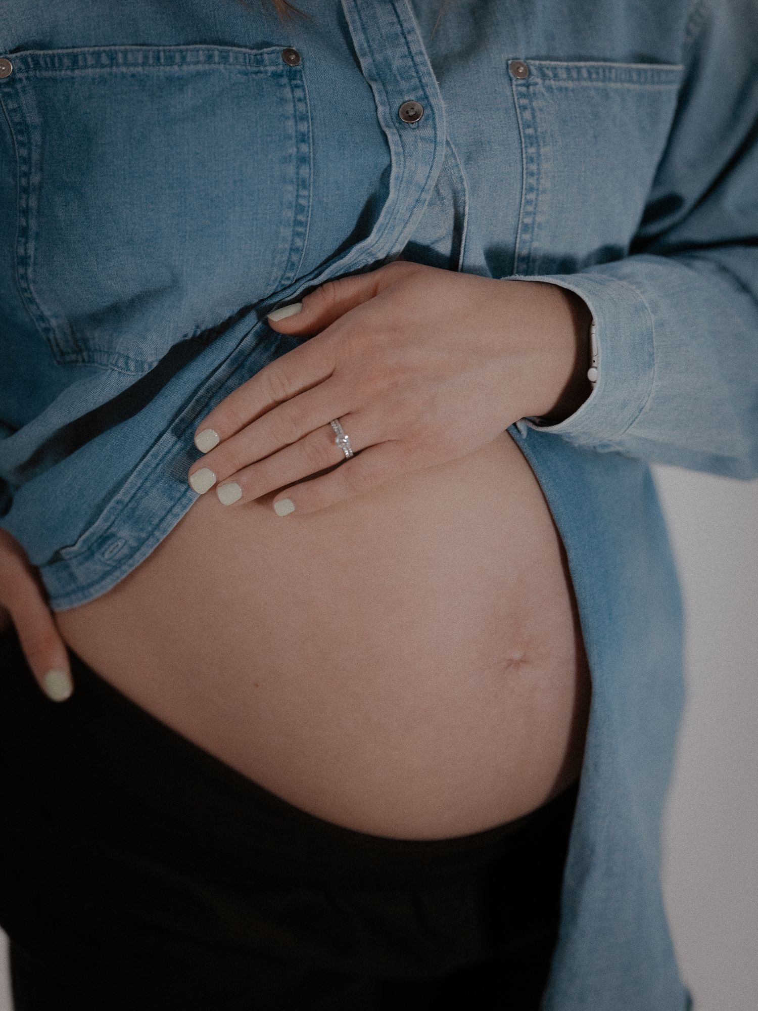 Close-up of pregnant woman in denim shirt cradling baby bump wearing a diamond engagement ring.