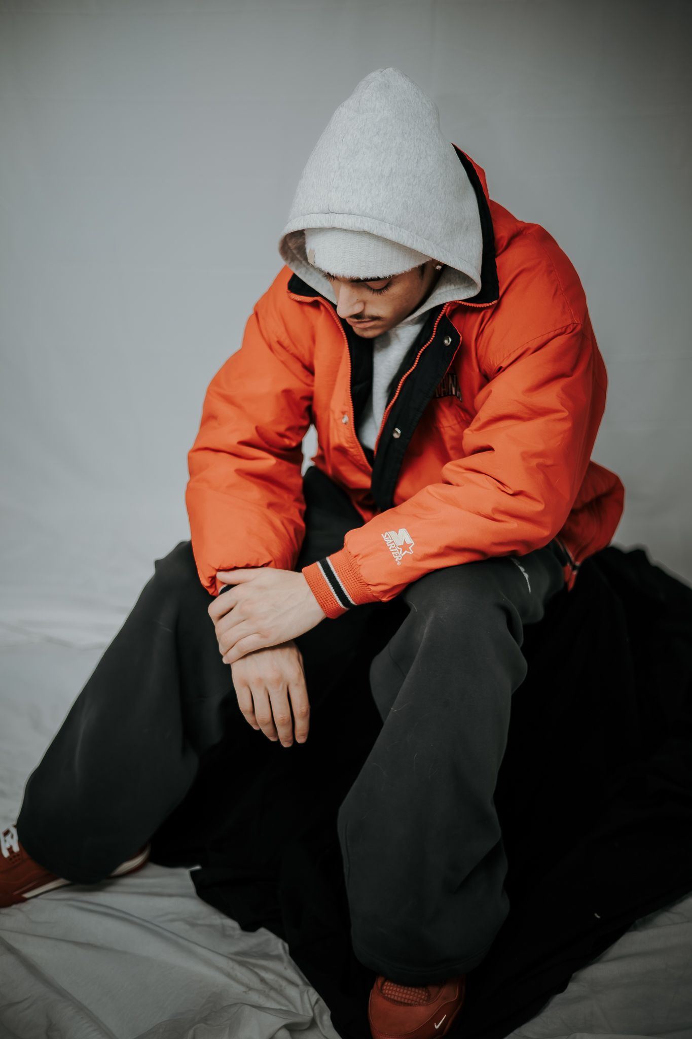 Young man in orange puffer jacket and gray hoodie sitting pensively against white background.