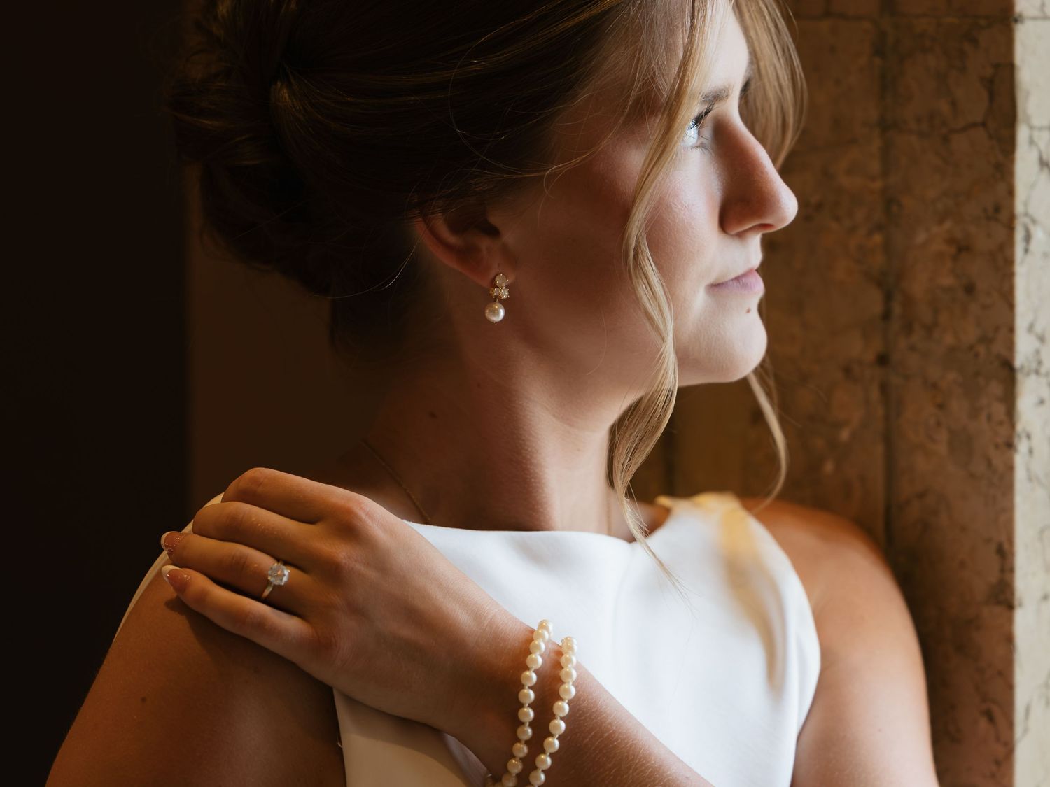 Bride wearing pearl bracelet and earrings with engagement ring gazing out a window.
