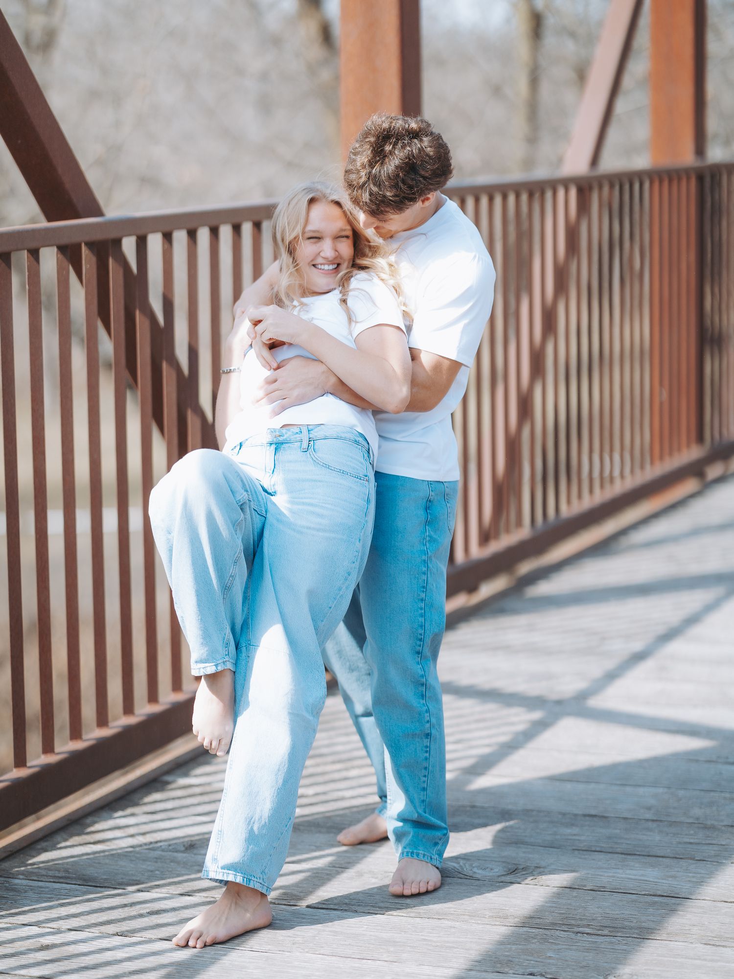 A couple embraces and smiles on a wooden bridge, both wearing white shirts and light blue jeans on a sunny day.