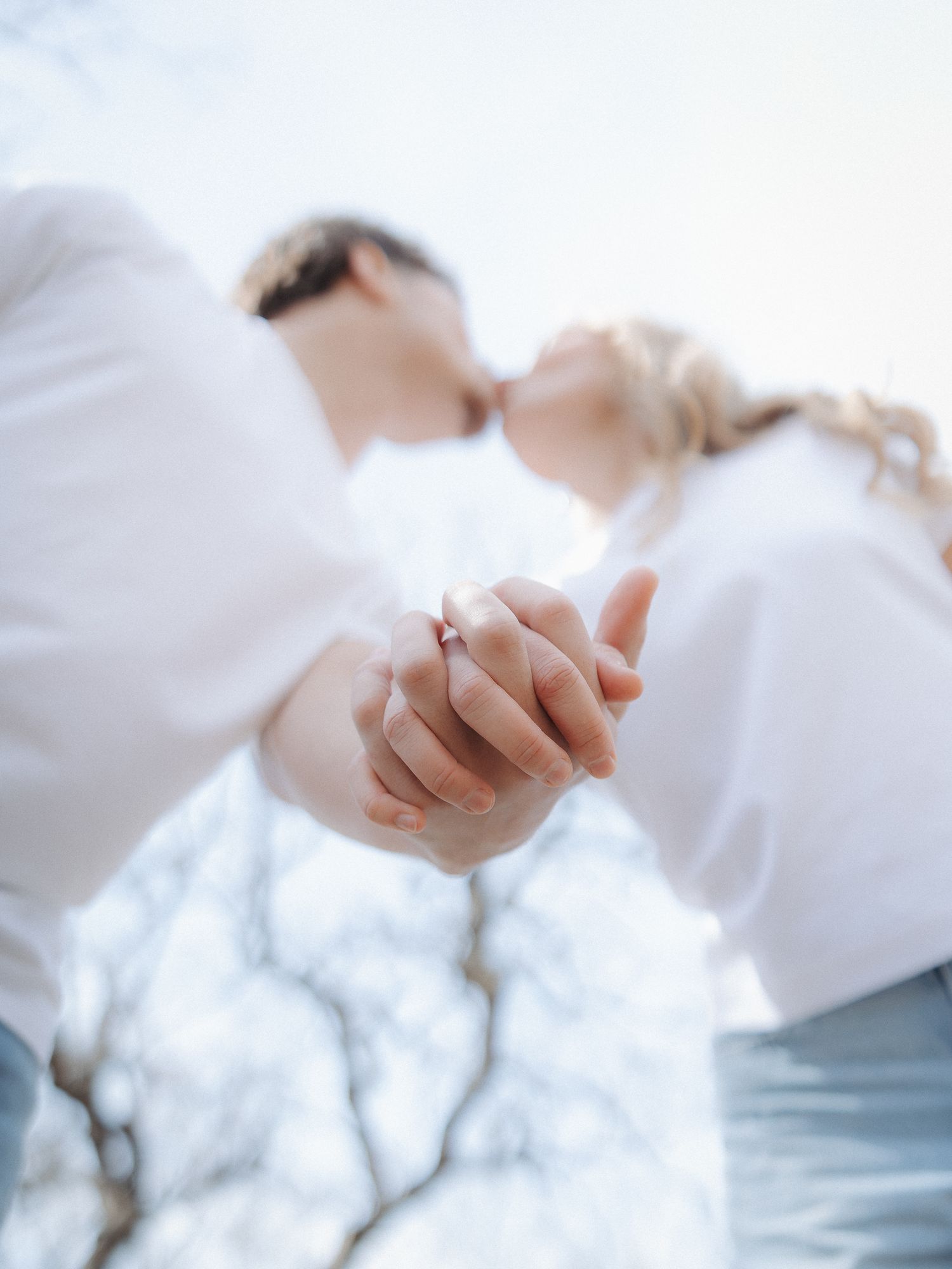 Couple kissing while holding hands with engagement ring visible, shot from below against bright sky.