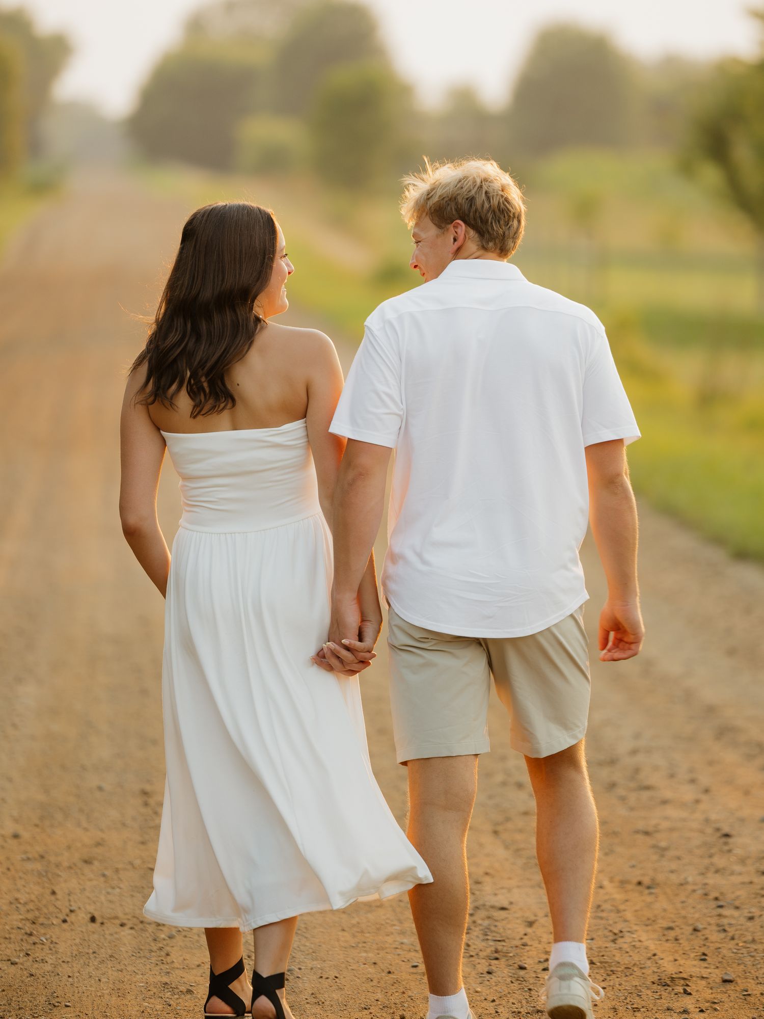 Couple holding hands walking down a dirt road at sunset, both dressed in white outfits.