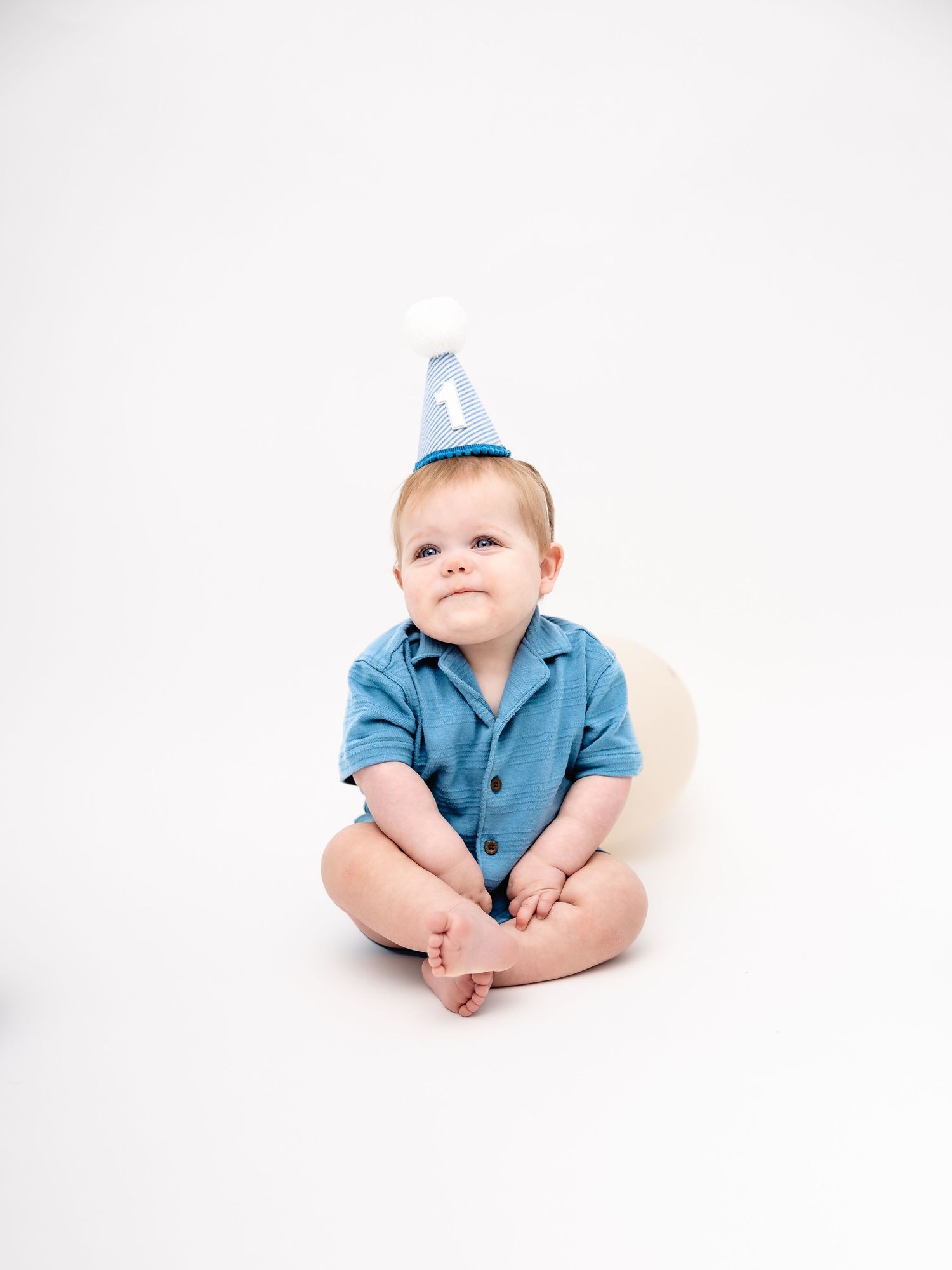 A baby boy in a blue outfit and birthday party hat sits on a white background with a balloon.