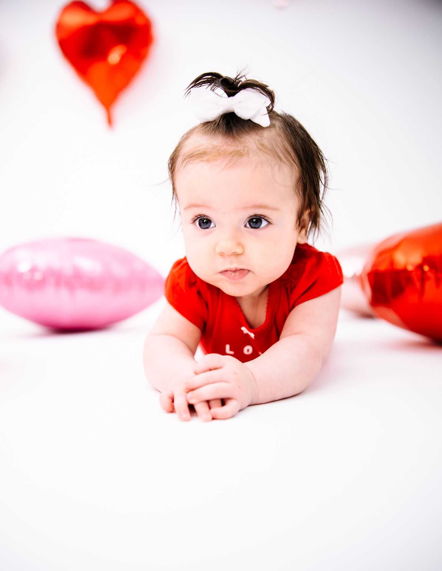 Adorable baby in red onesie lying on white floor surrounded by heart-shaped balloons.