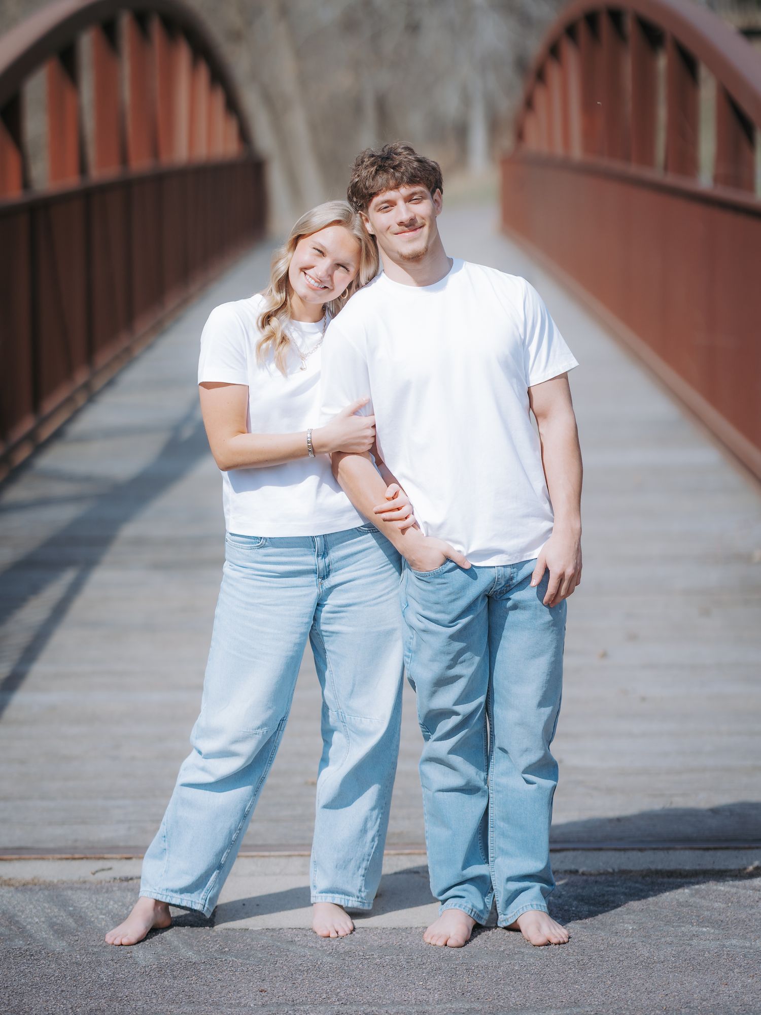 A smiling couple in matching white tees and light jeans pose barefoot on a red bridge outdoors.