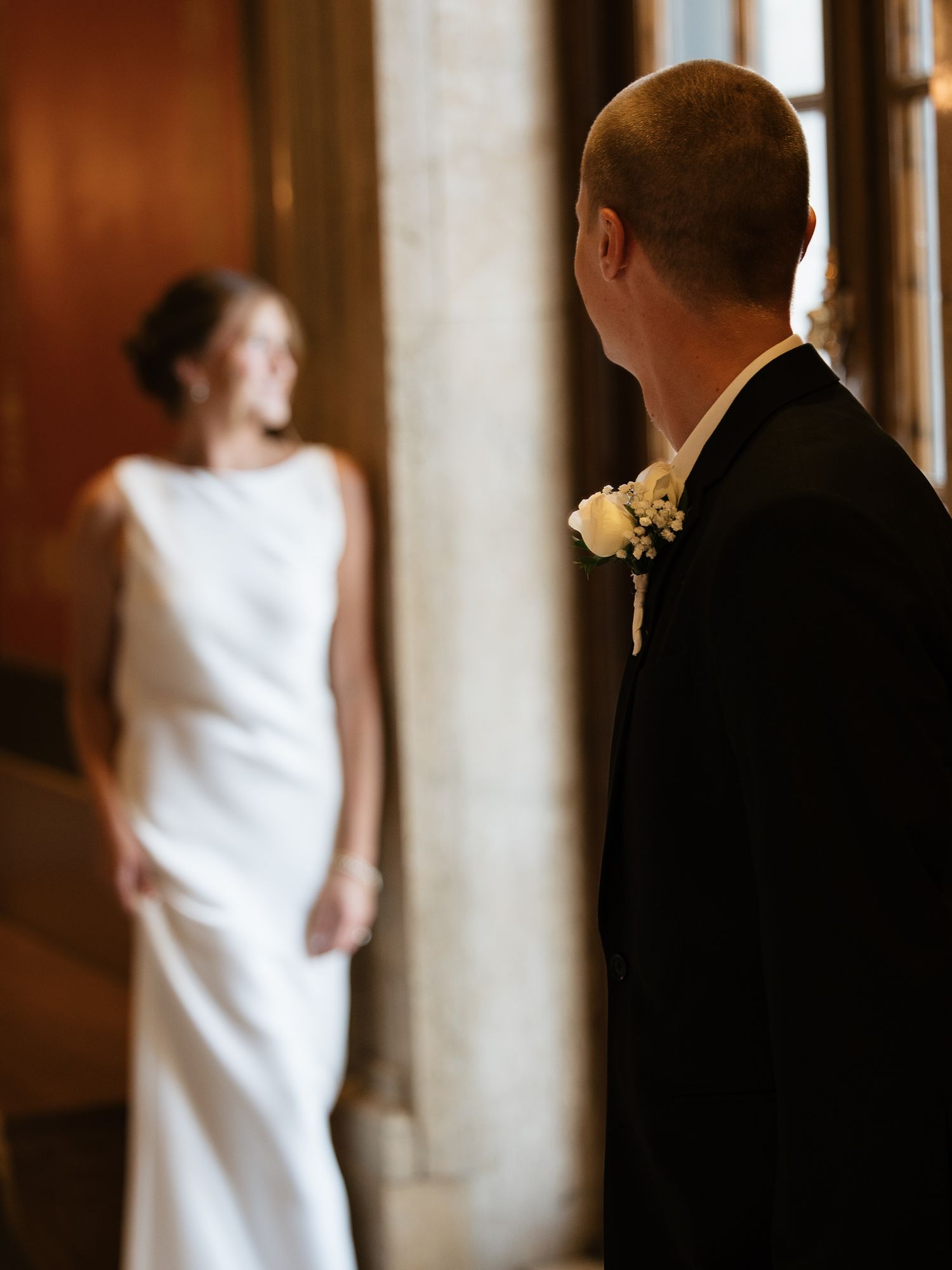 Groom in black suit watching bride in white dress standing by window.