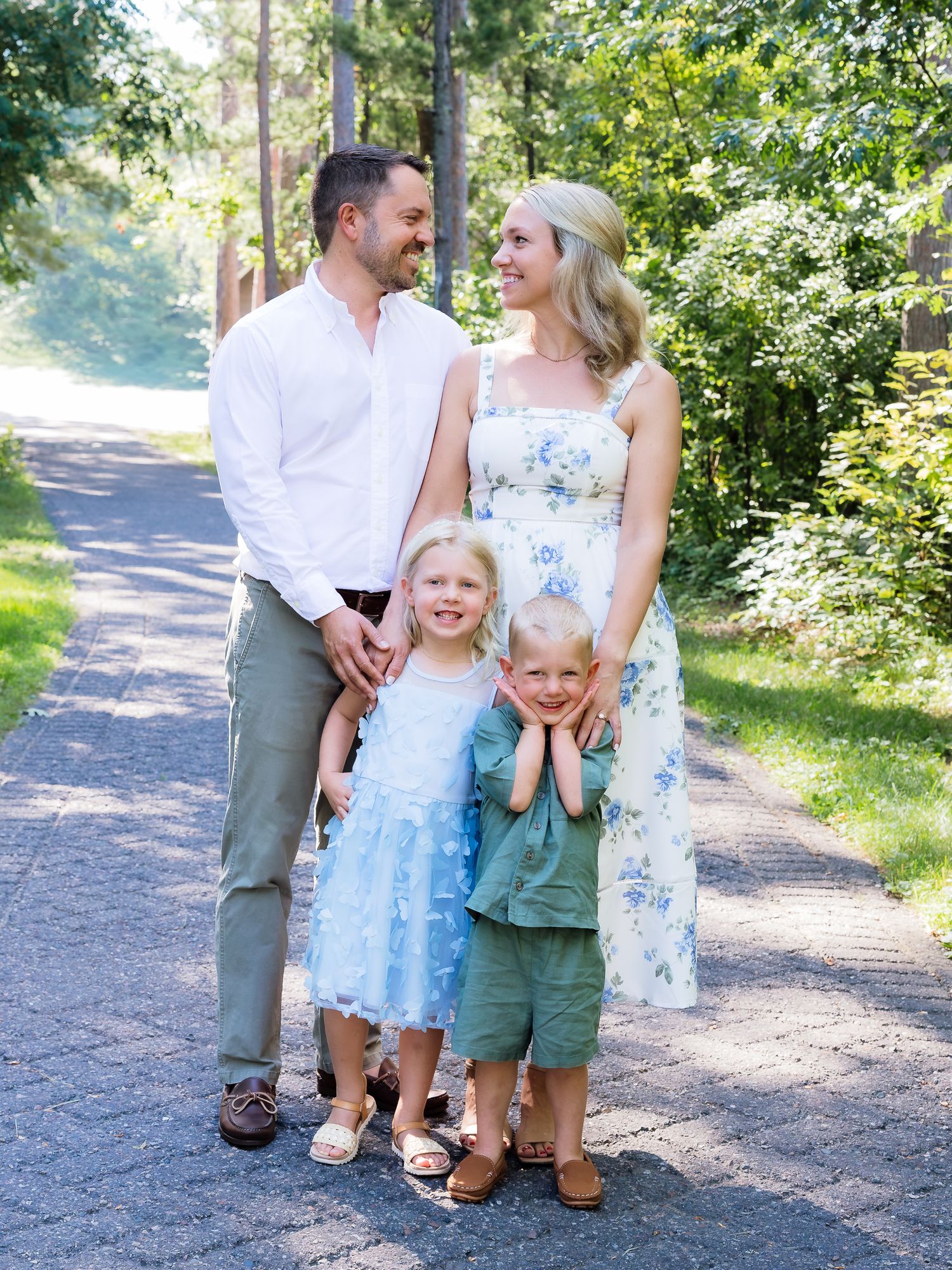 A happy family of four poses on a sunny tree-lined path, parents smiling at each other lovingly.