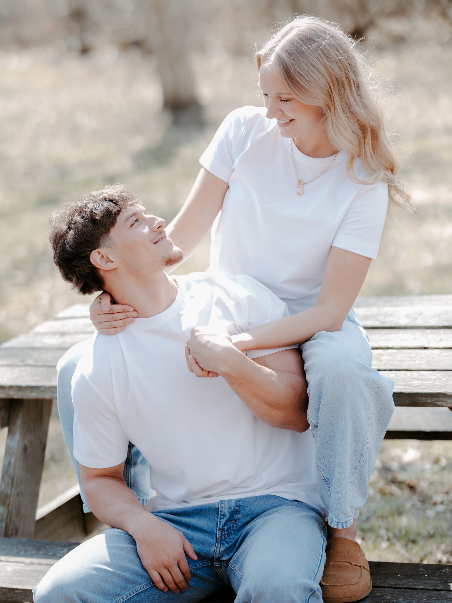 A young couple in matching white t-shirts and jeans share a tender moment on a wooden bench outdoors.