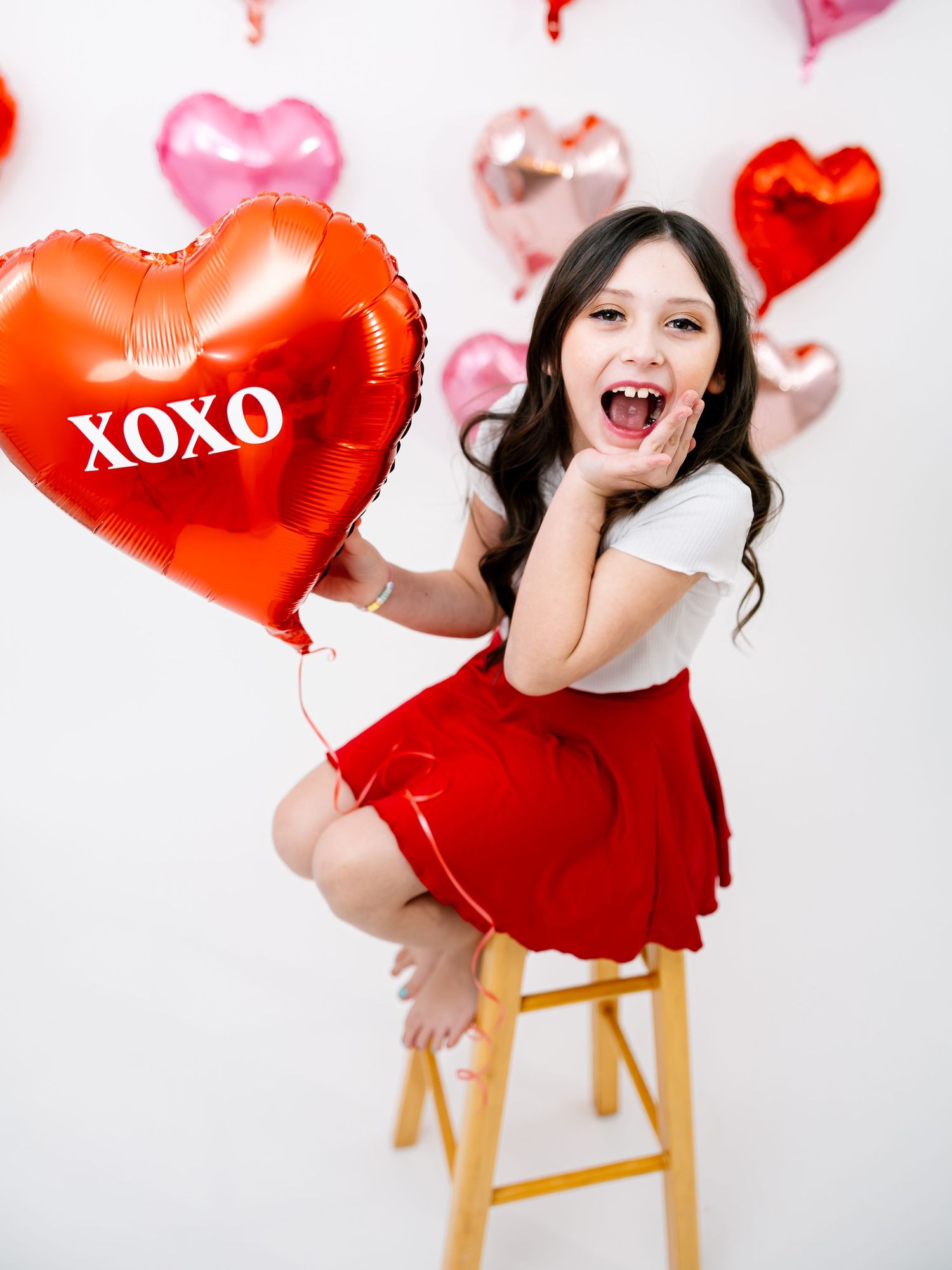 Excited girl in red skirt holding XOXO heart balloon sitting on stool with Valentine heart balloons in background.