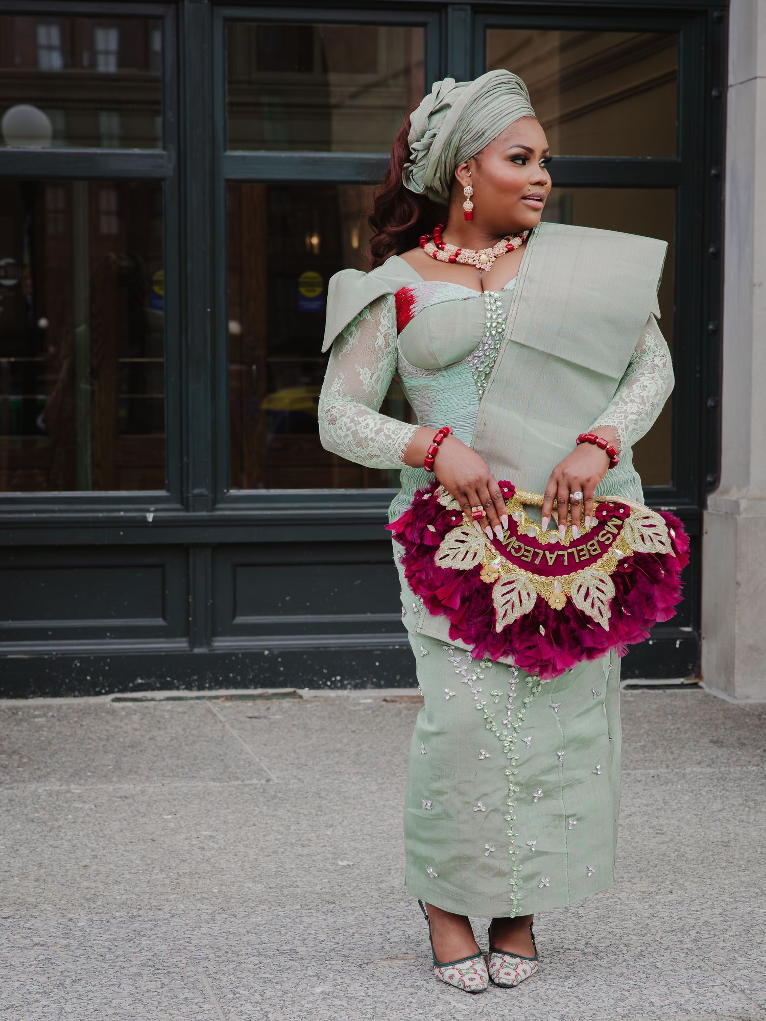 A woman in an elegant mint green Nigerian traditional outfit with a floral clutch poses outdoors.