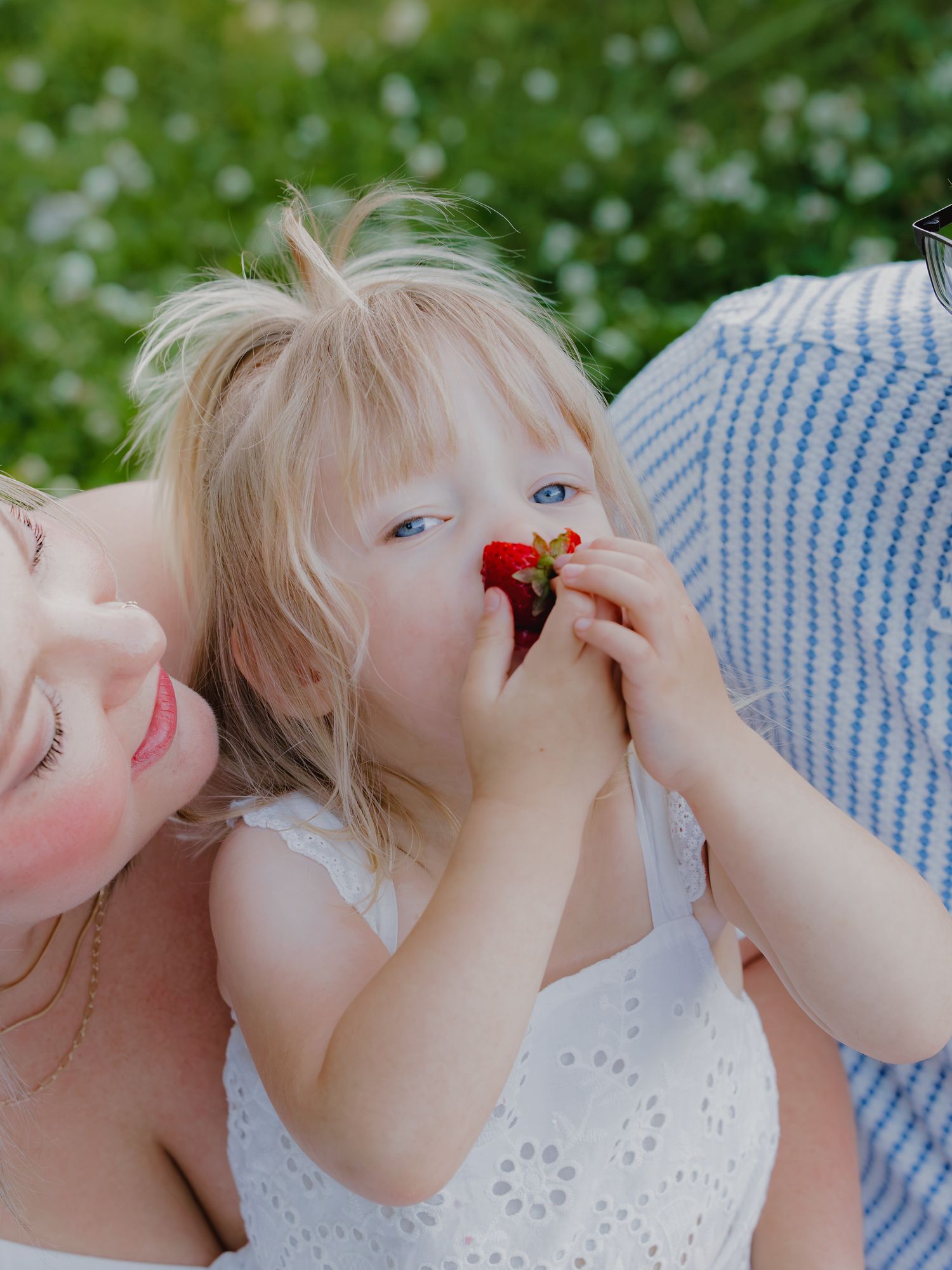 A young blonde toddler eats a strawberry while sitting with a woman in a white dress outdoors.