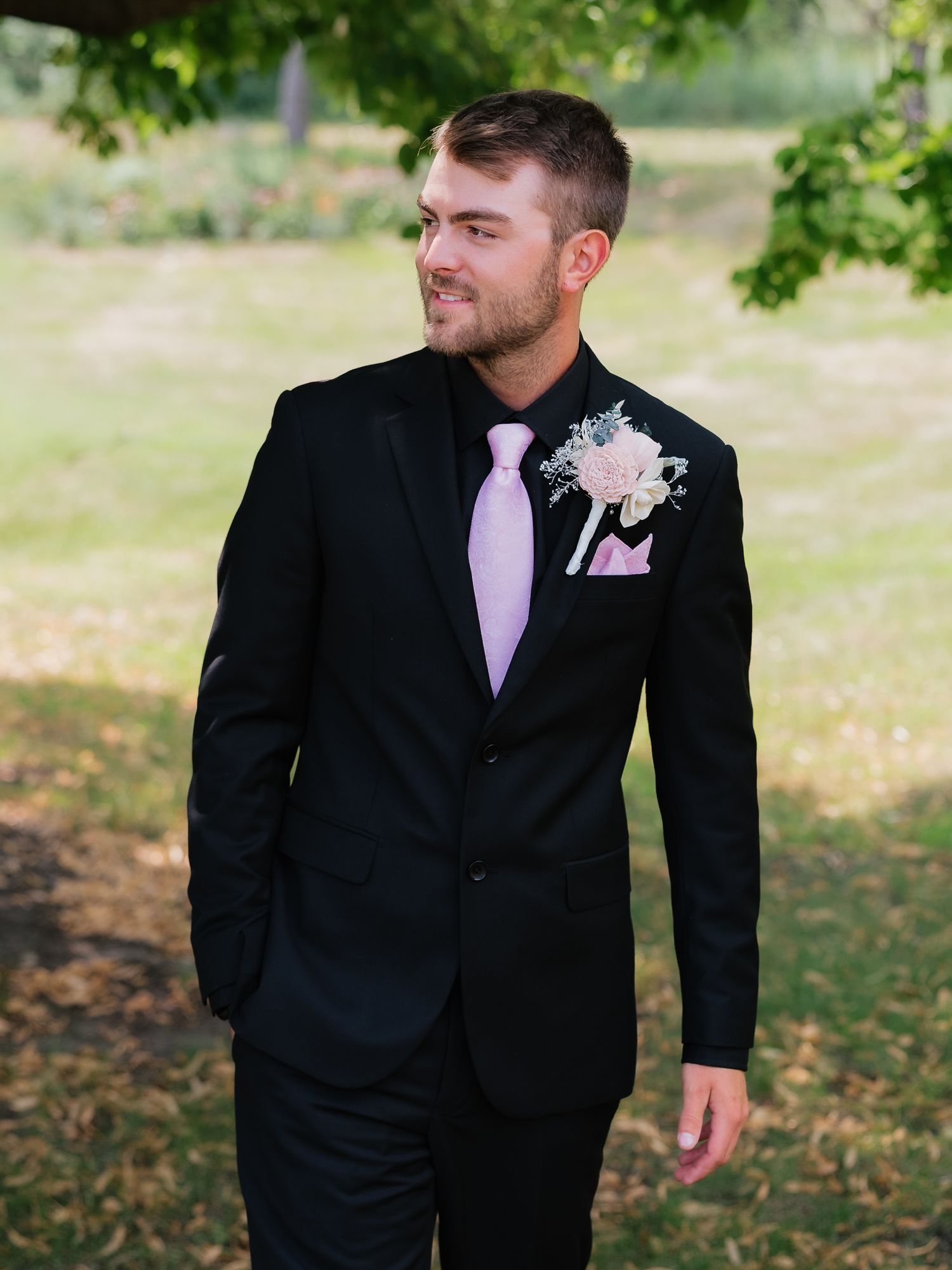 Groom in black suit with pink tie and boutonniere standing outdoors.