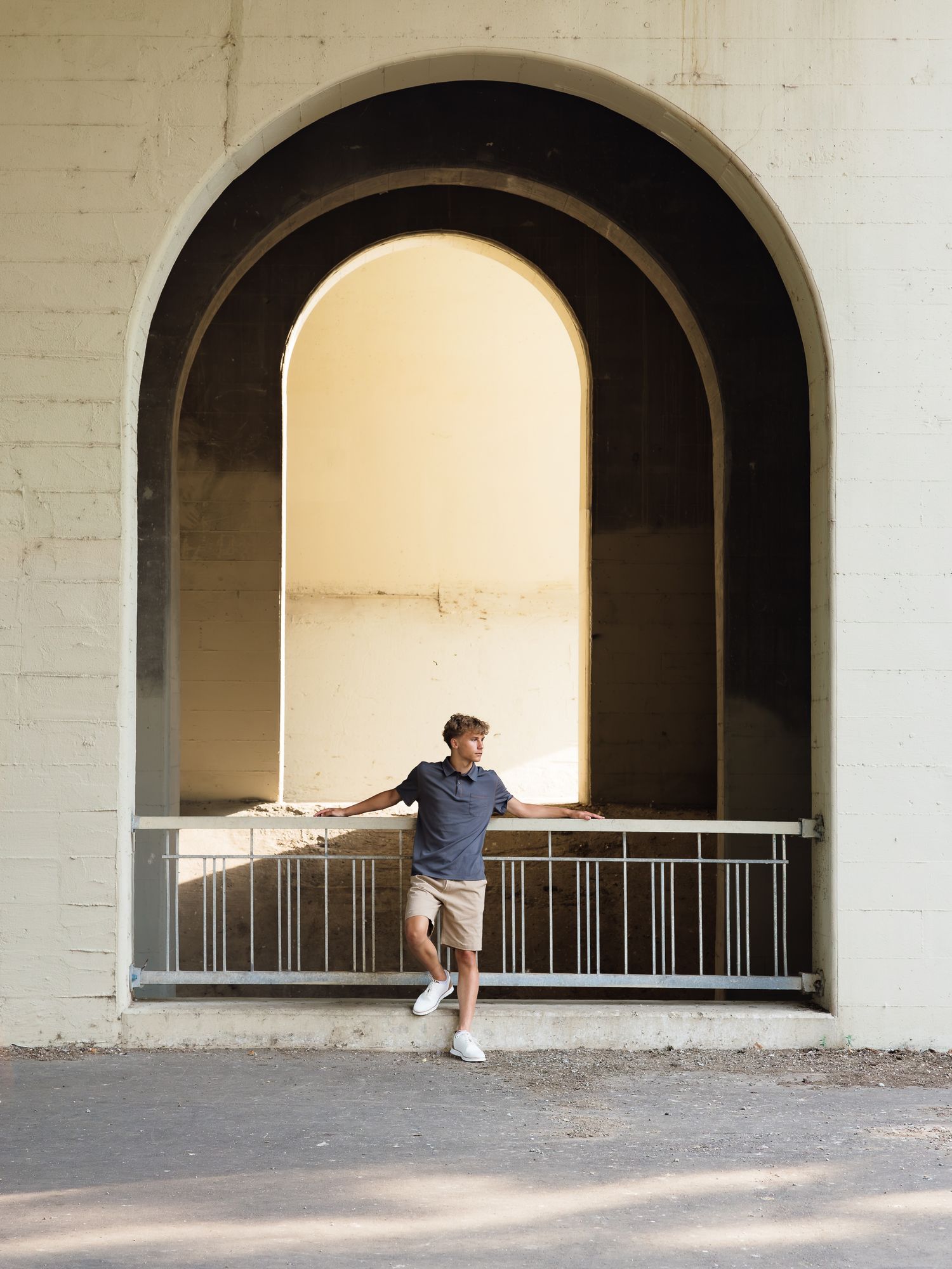 Young man leaning on a metal railing inside a large arched tunnel entrance with white brick walls.