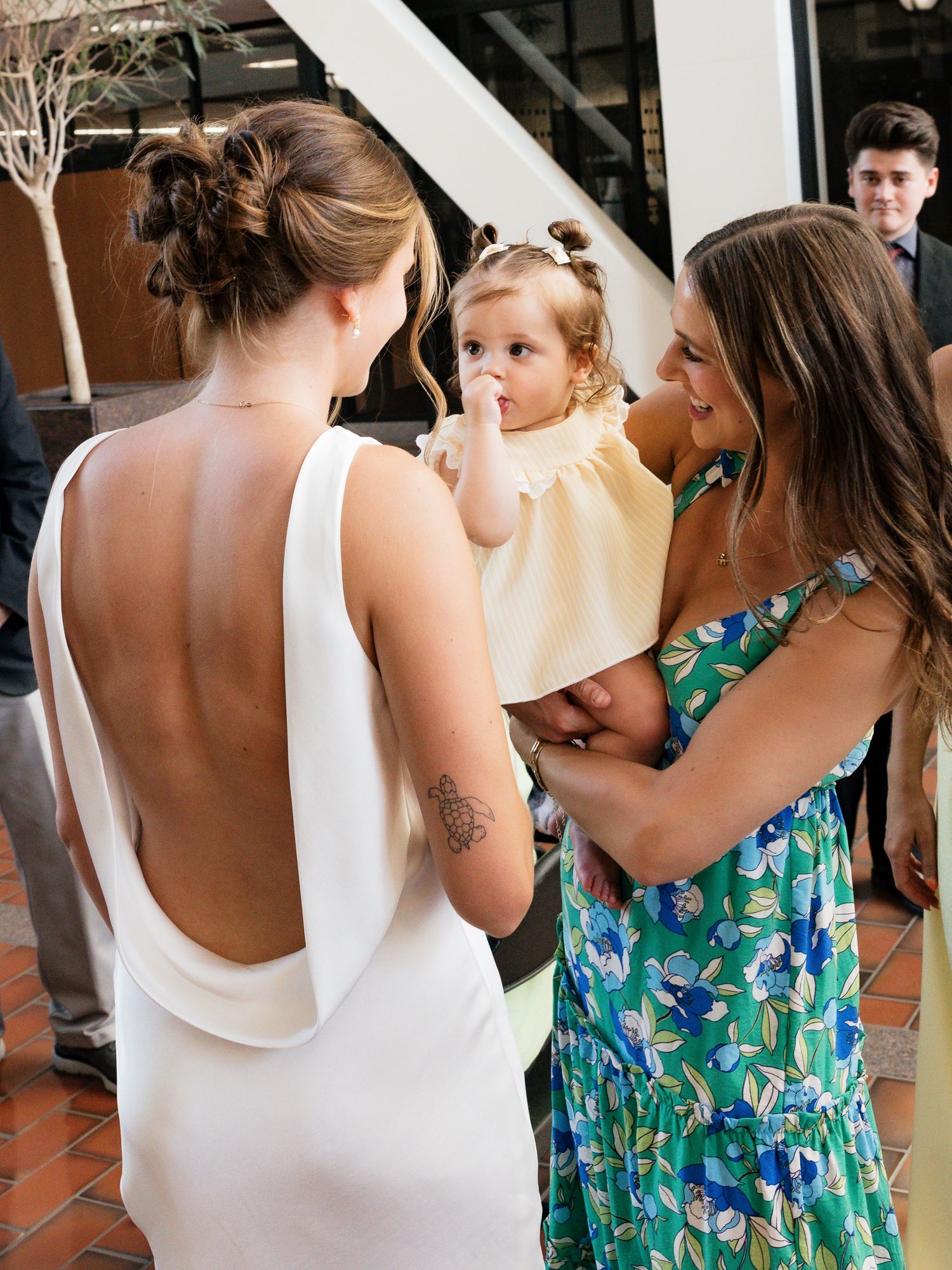 Bride in white backless dress with tattoo talks to woman in green floral dress holding a toddler girl.