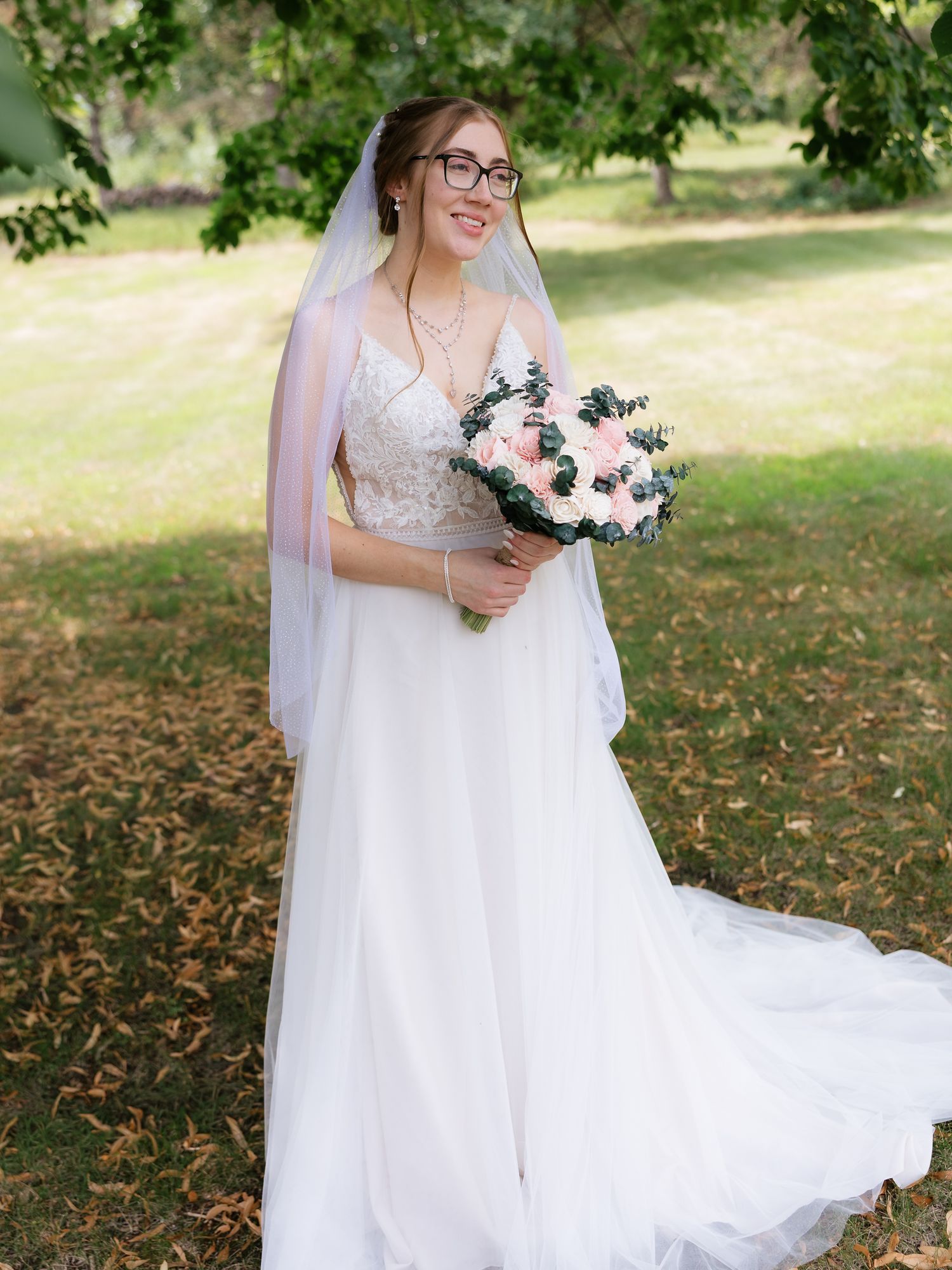 Smiling bride in white lace gown and veil holding pink floral bouquet outdoors.