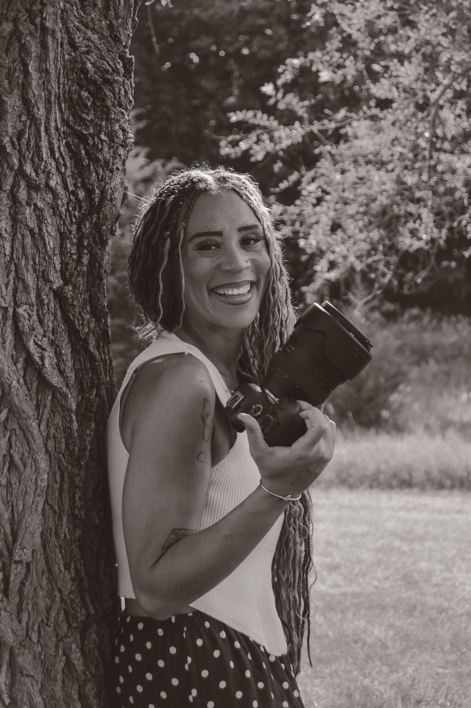 Smiling woman with long braids leans against tree holding camera in black and white photo.