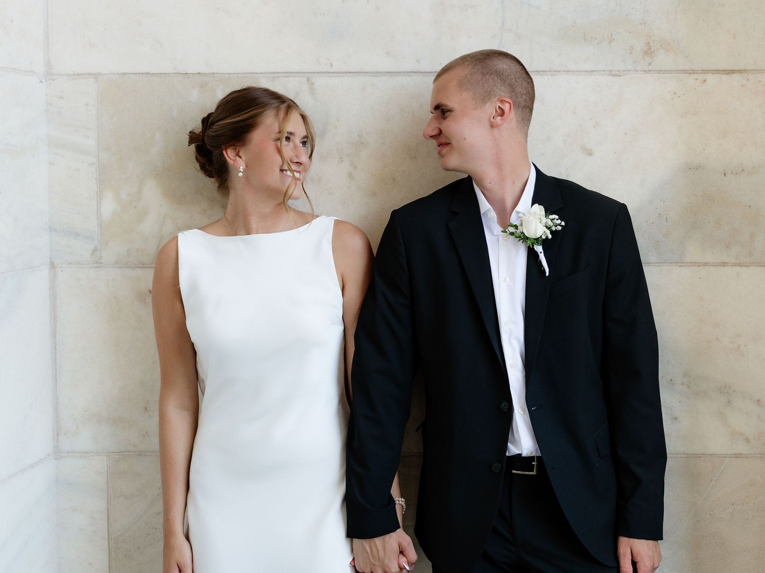 Smiling bride and groom holding hands in front of a marble wall on their wedding day.