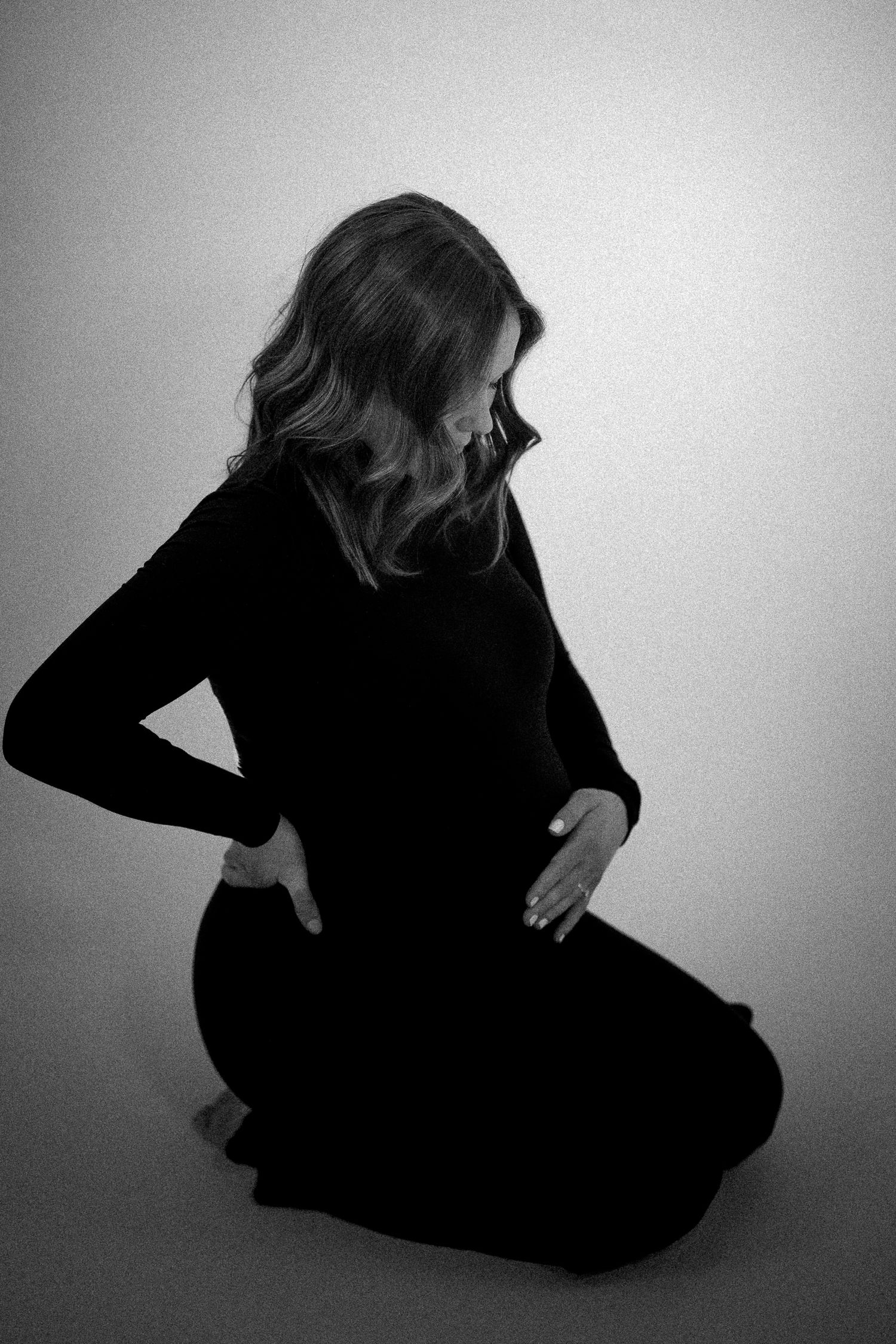Black and white maternity photo of pregnant woman in black dress sitting on floor cradling baby bump.