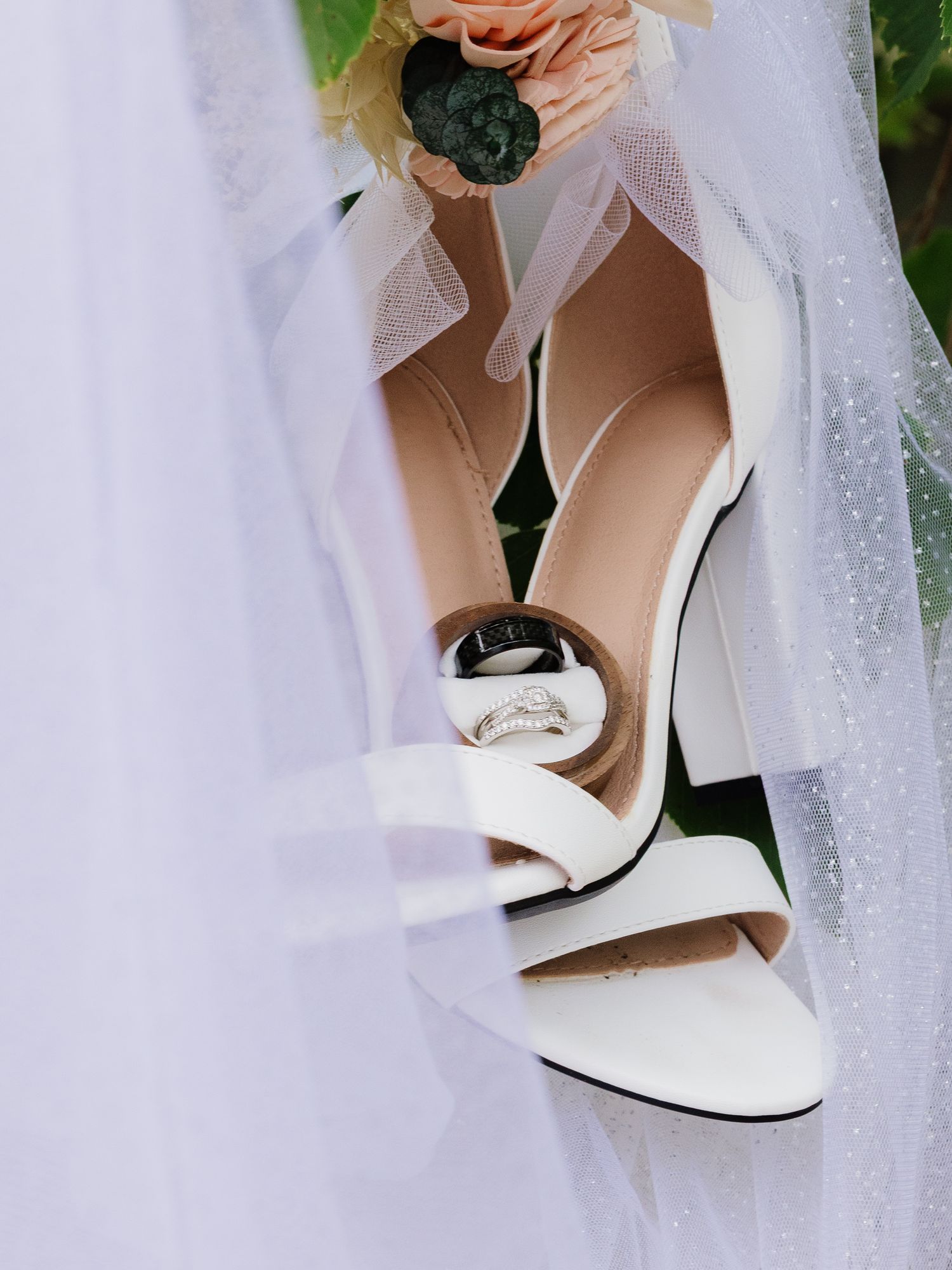 White block heel wedding shoes with diamond rings resting inside heel near bridal veil.