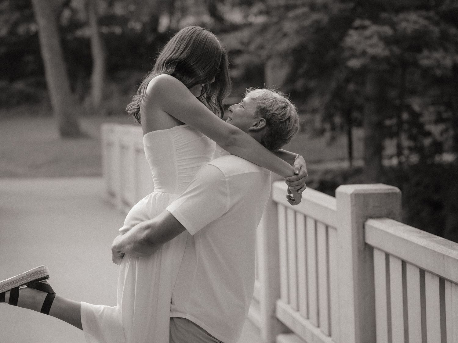 Black and white photo of a couple embracing romantically on a wooden bridge, woman in white dress.