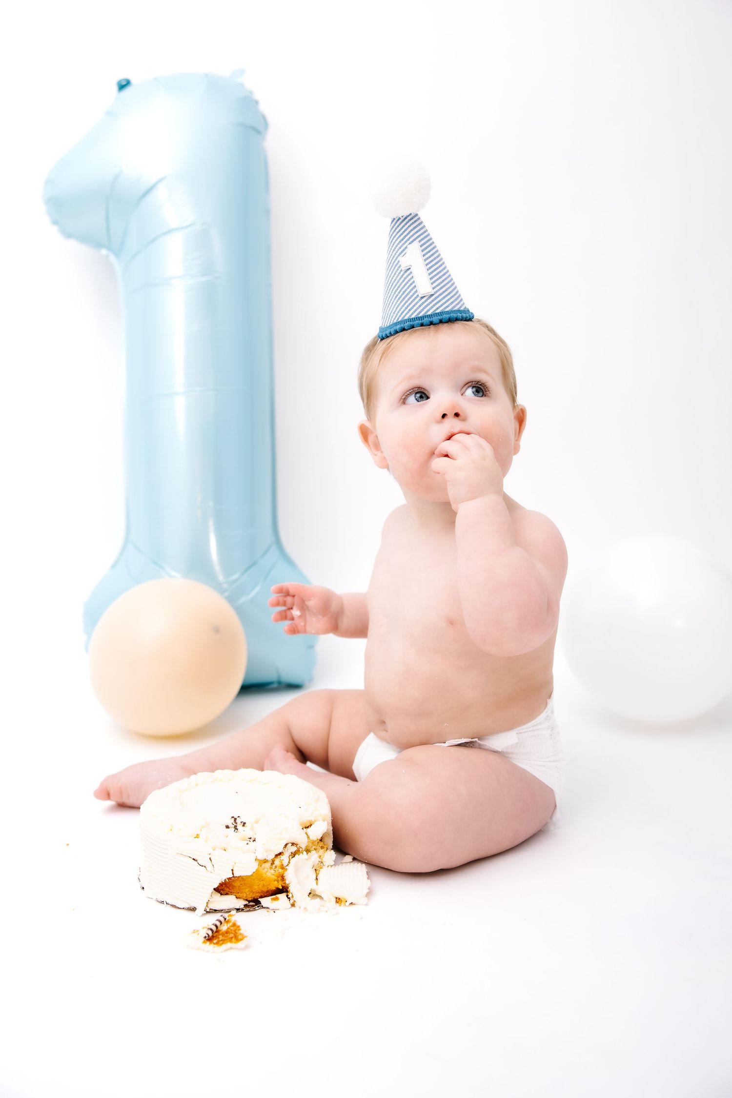 Baby in diaper and party hat celebrating first birthday with smashed cake and blue number one balloon.