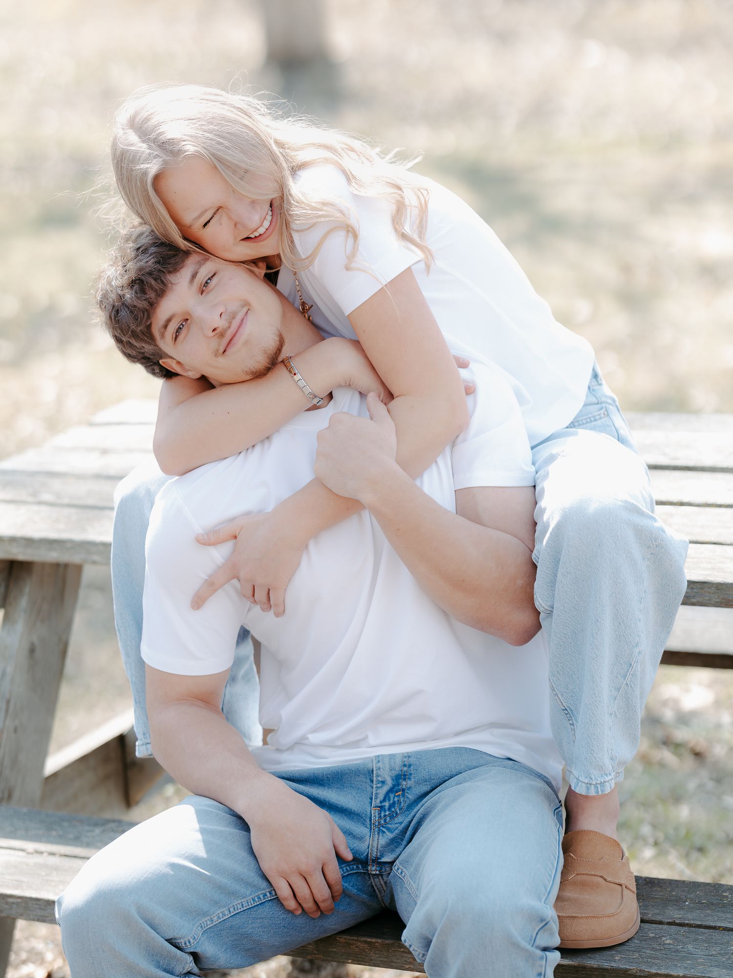 Playful couple posing on a wooden bench outdoors with woman hugging man from behind.