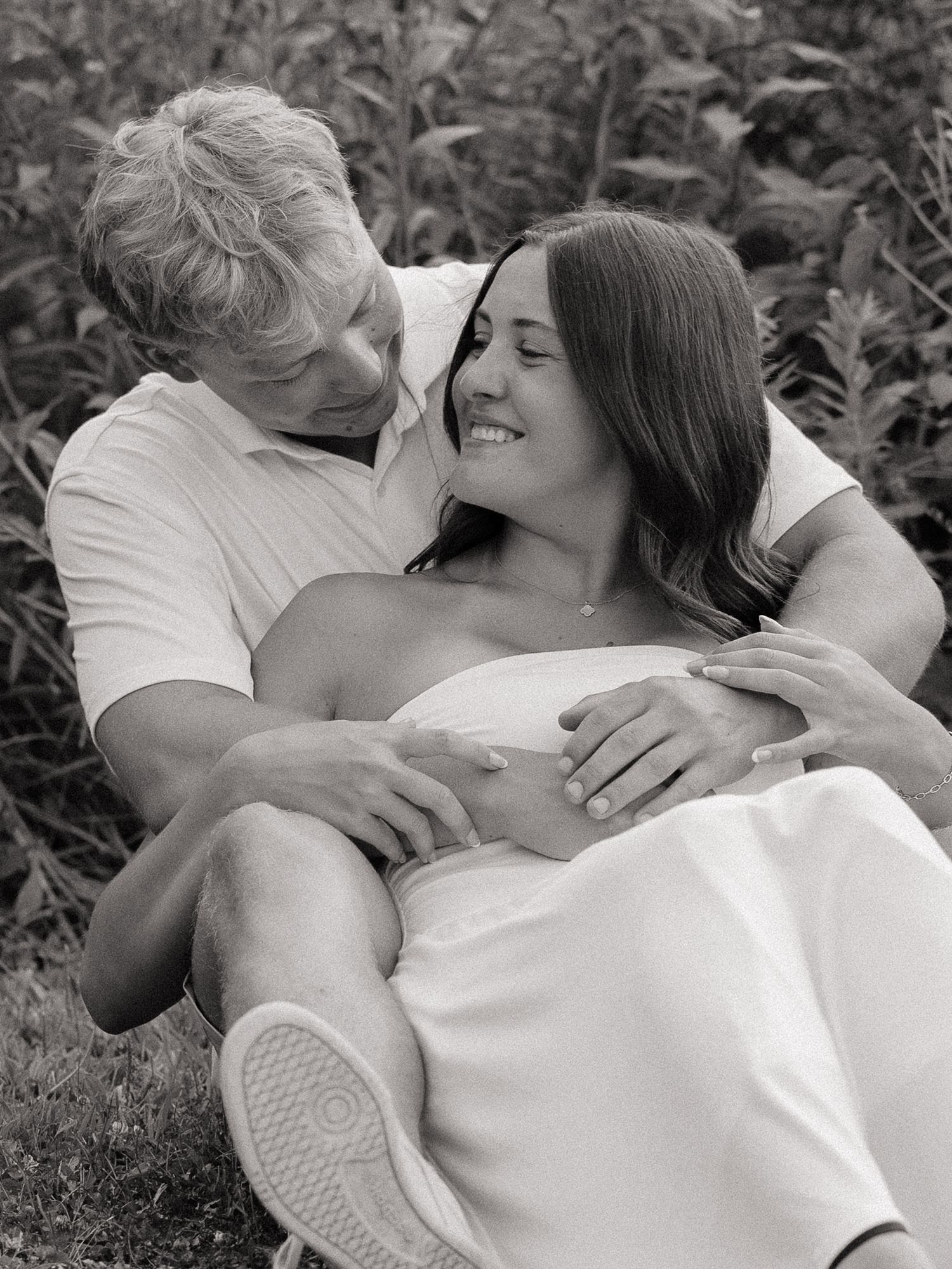 Black and white photo of a smiling couple sitting together in a lush garden, sharing a tender romantic moment.