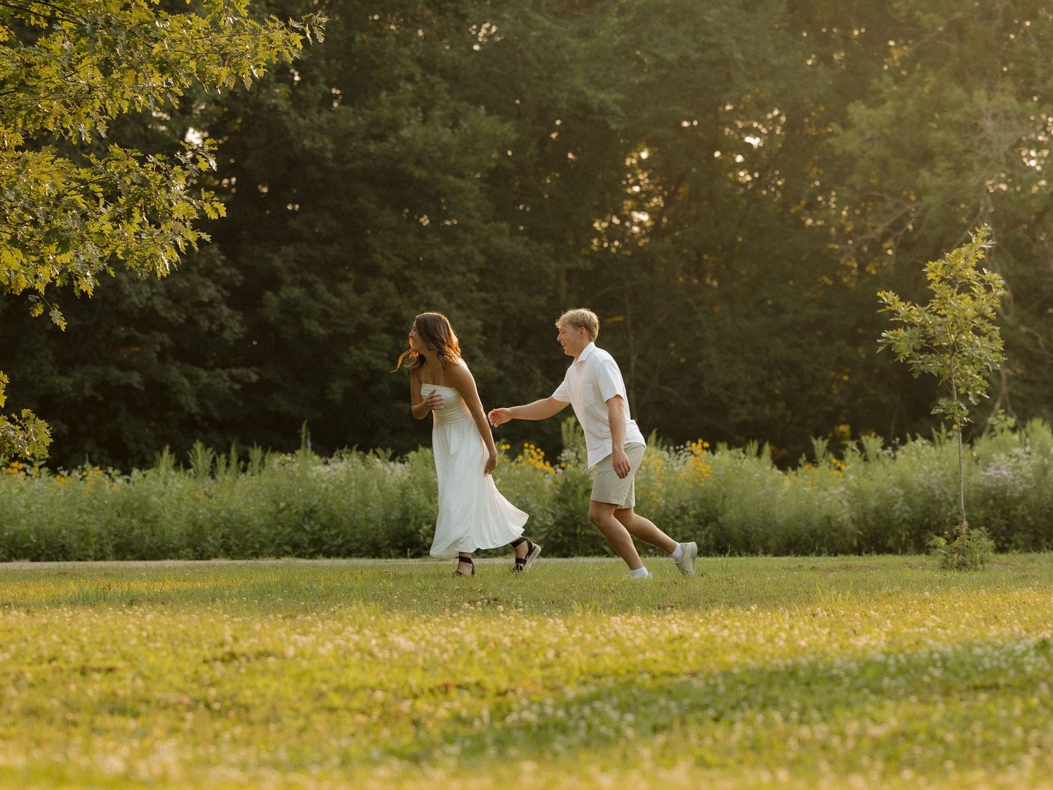 Couple holding hands and dancing in a sunlit green park surrounded by lush trees during golden hour.