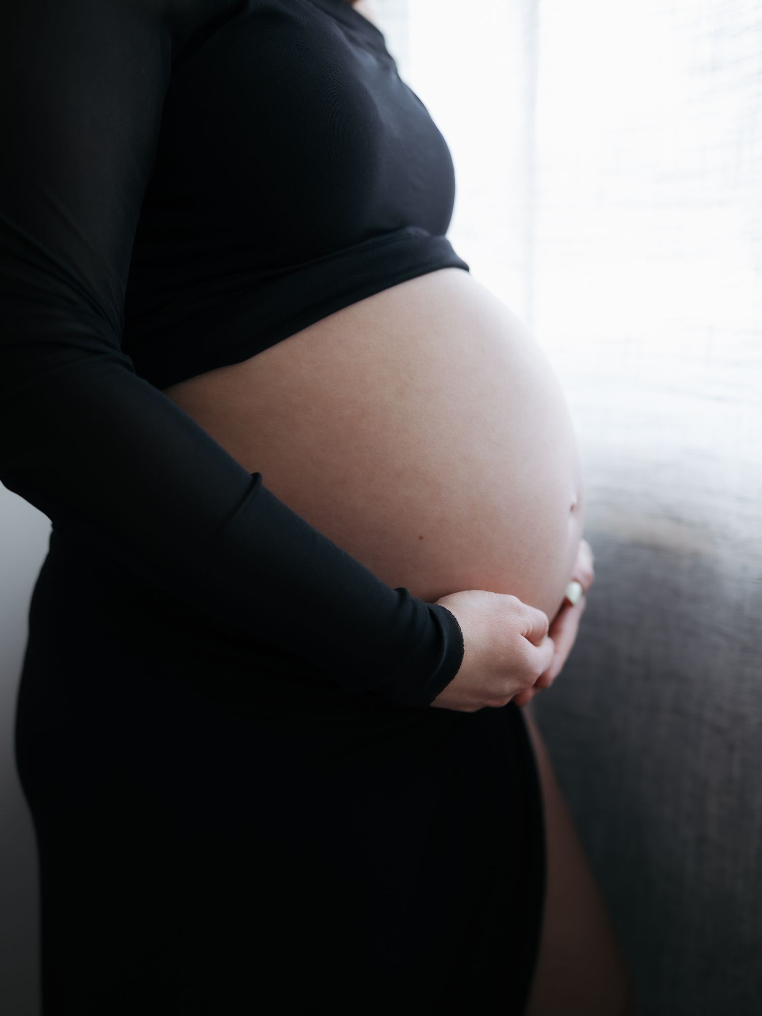Pregnant woman in black crop top and skirt cradling her bare baby bump near a bright window.