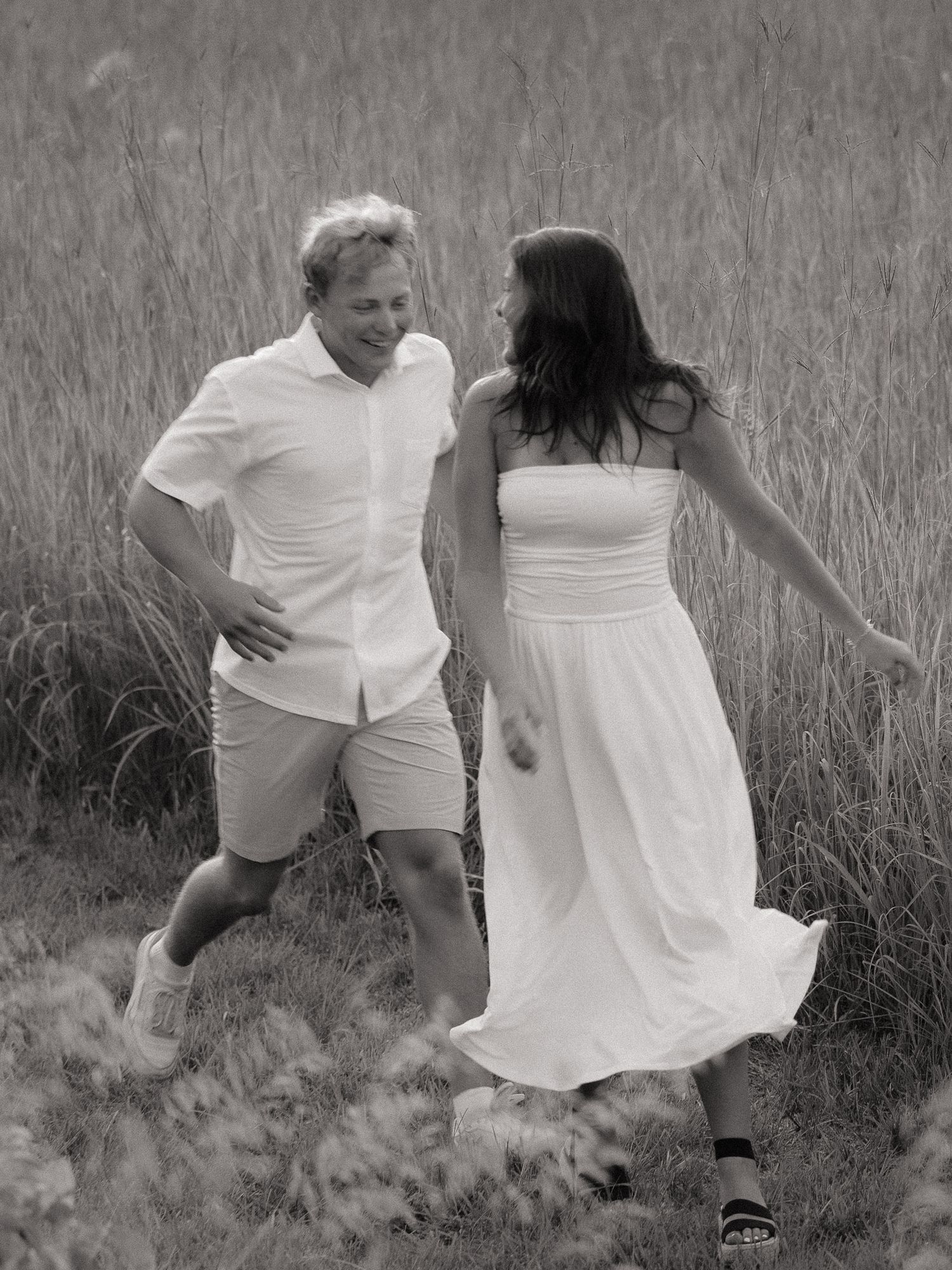 Black and white photo of a couple laughing and walking through tall grass, wearing matching white outfits.