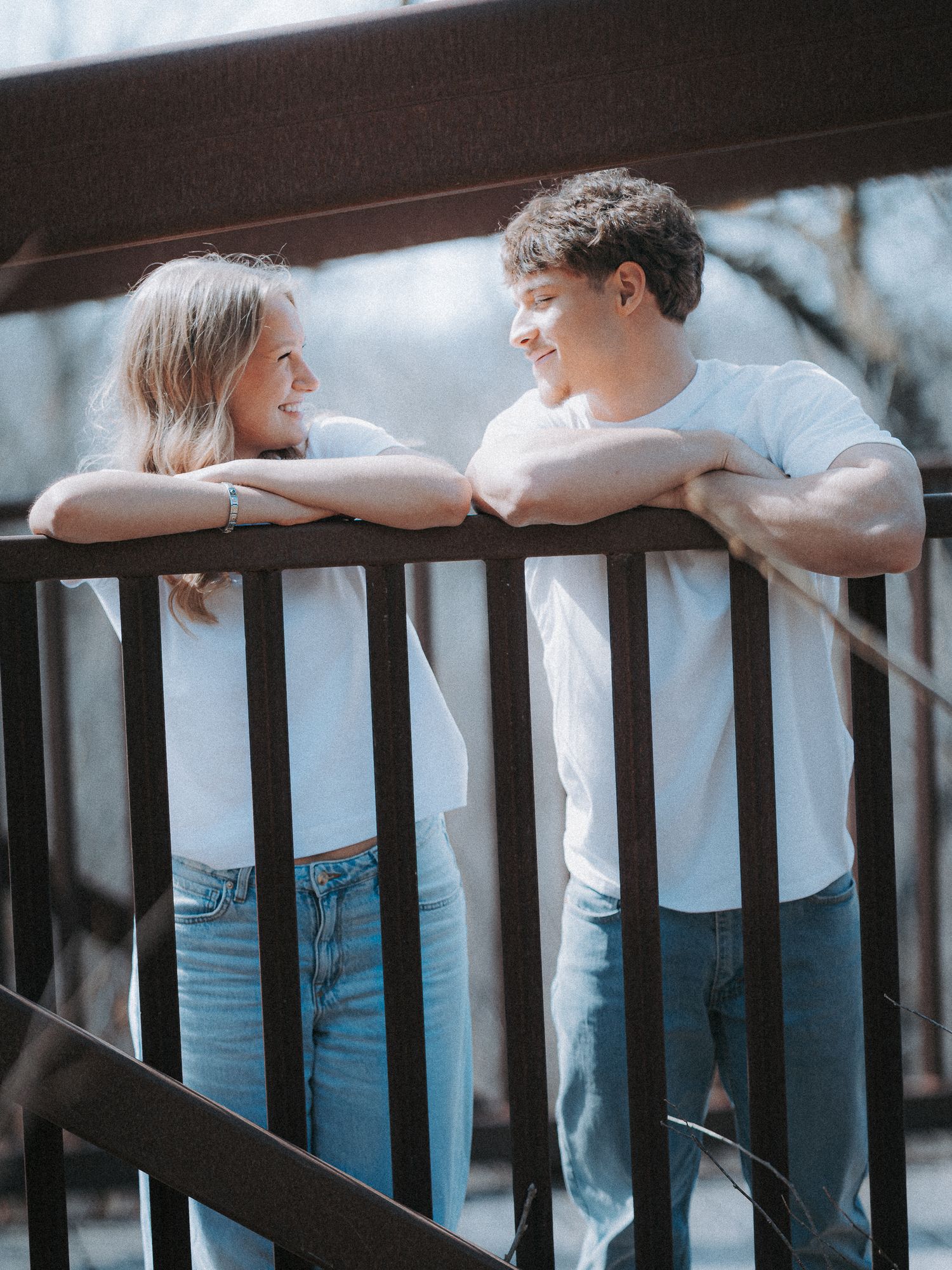 A young couple in matching white tees and jeans smile at each other while leaning on a railing outdoors.