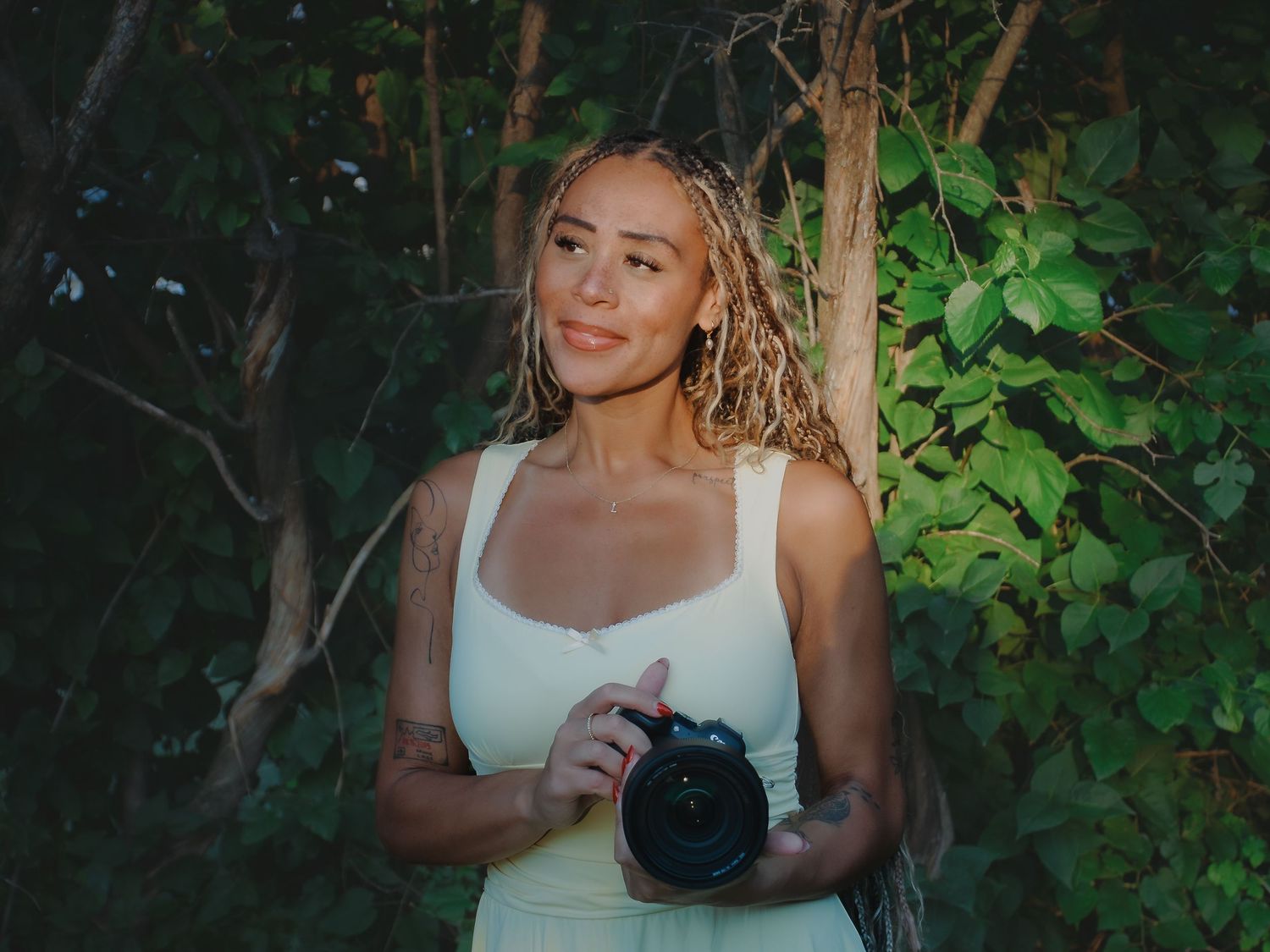 Young woman with blonde locs holding a camera, wearing a white dress, standing outdoors among green trees.