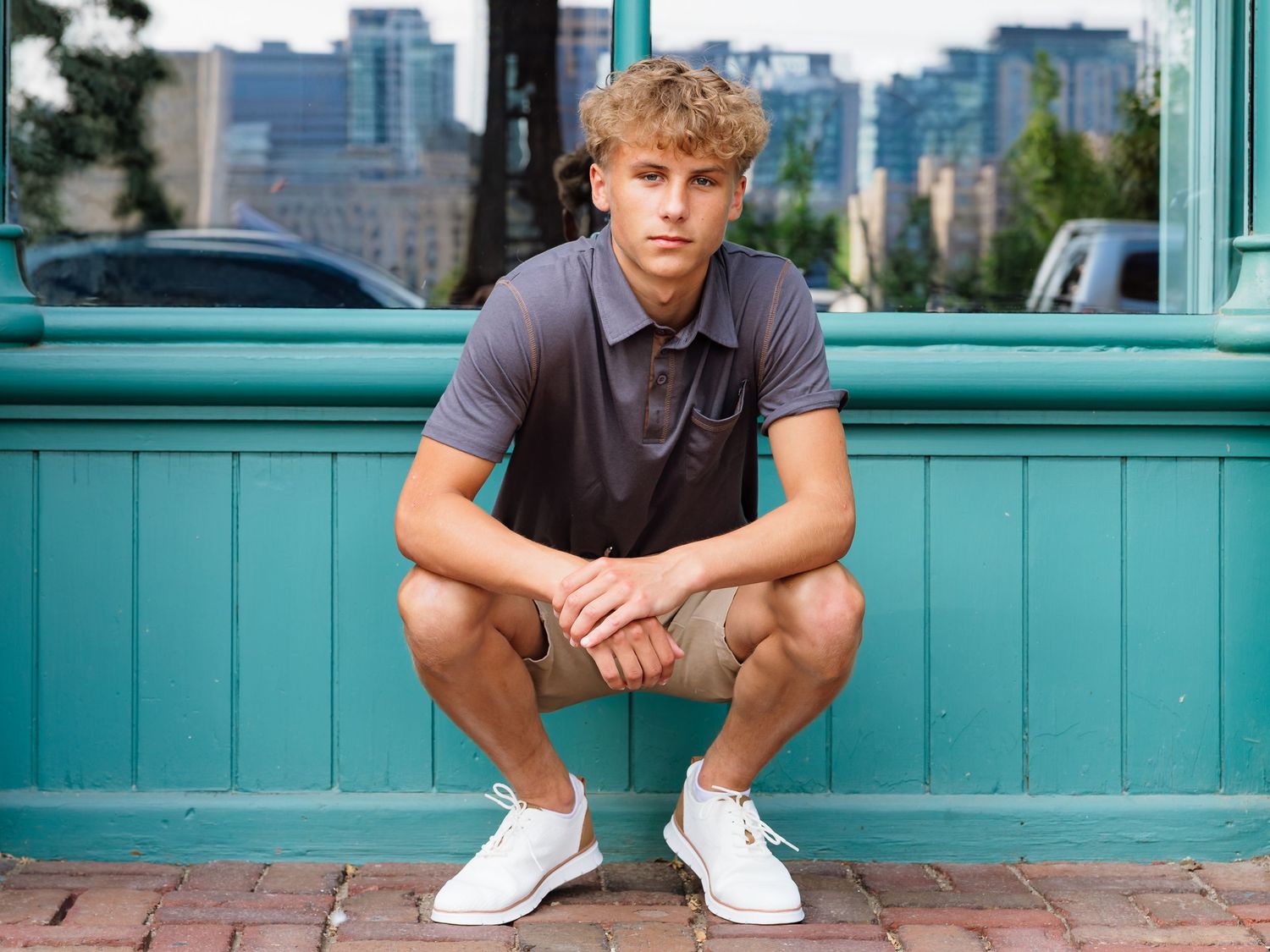 Confident young man in gray polo squatting in front of a teal wall in urban setting.