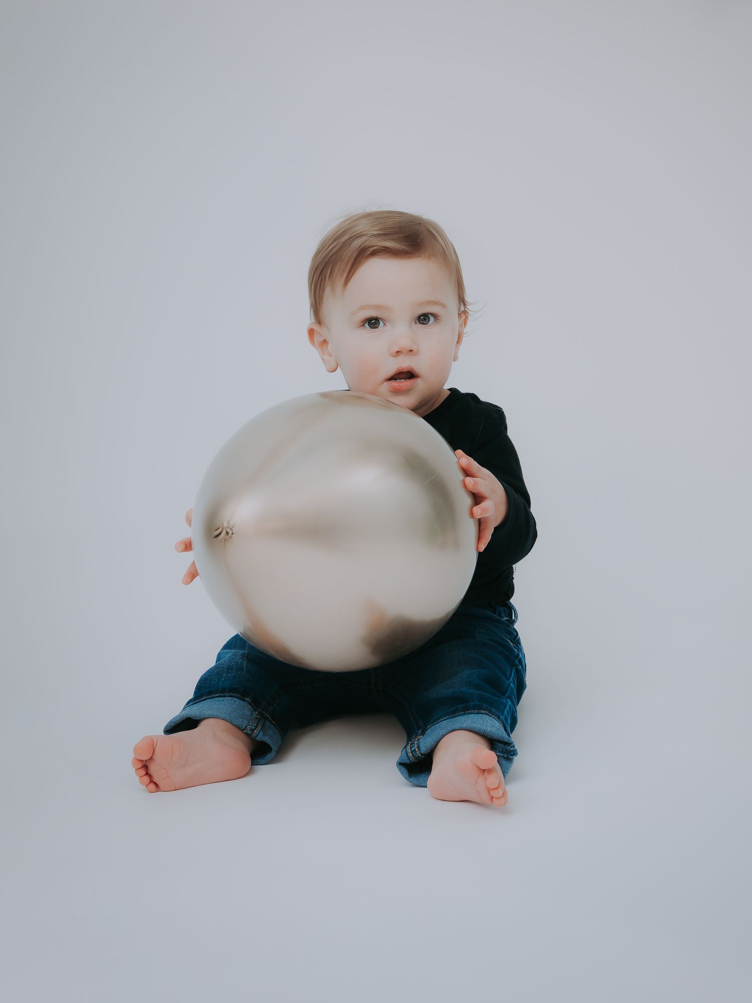Curious baby in black shirt sitting on white floor holding a large shiny silver metallic balloon.